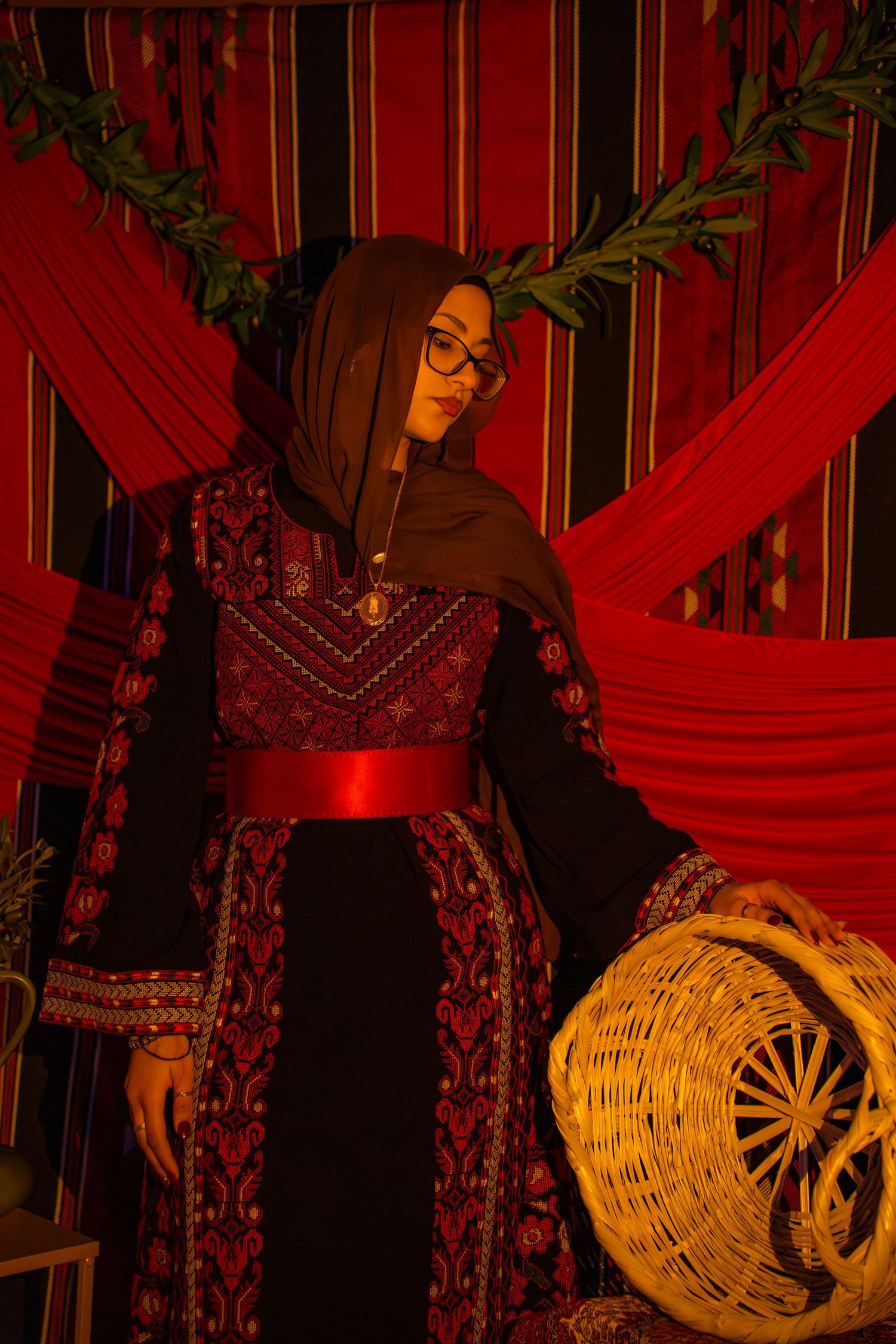 Traditional Middle Eastern attire with embroidered dress and red sash against striped backdrop with wicker basket.