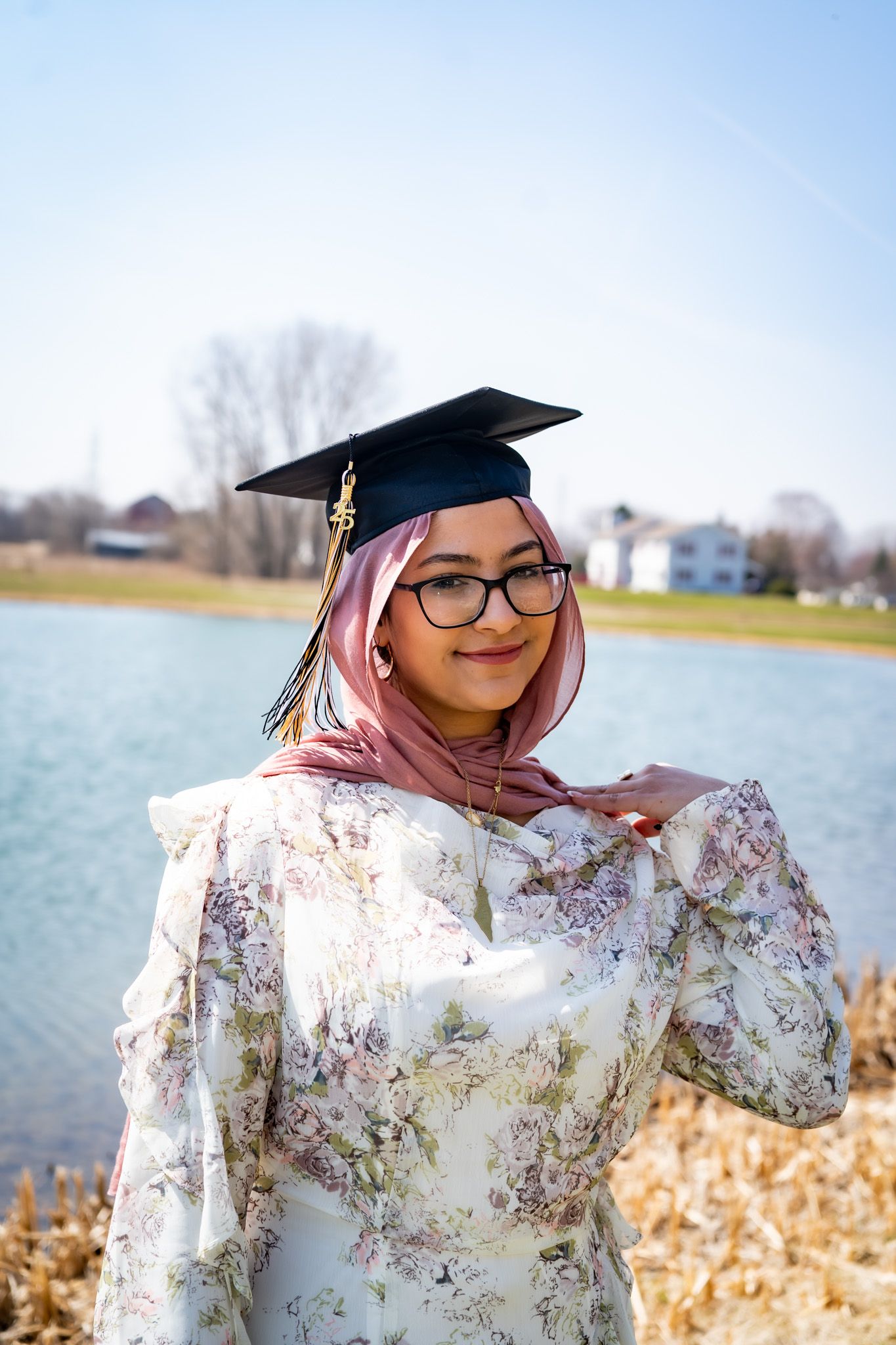 Graduate in floral dress and cap poses by waterfront on sunny day.