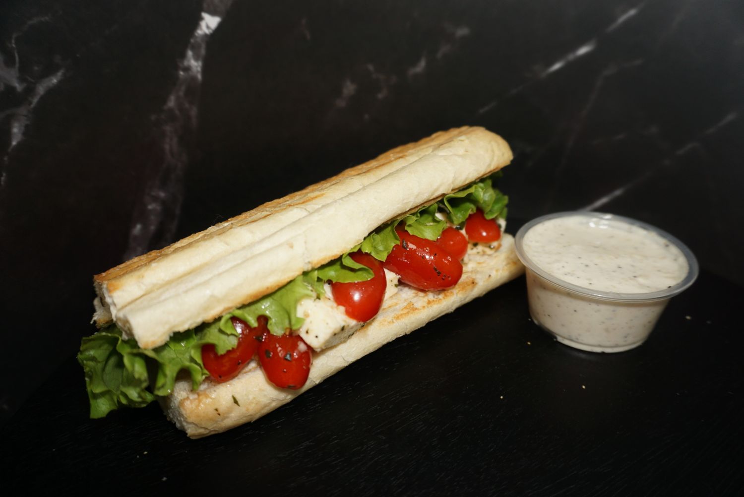 Fresh submarine sandwich with lettuce and tomatoes on dark marble surface.