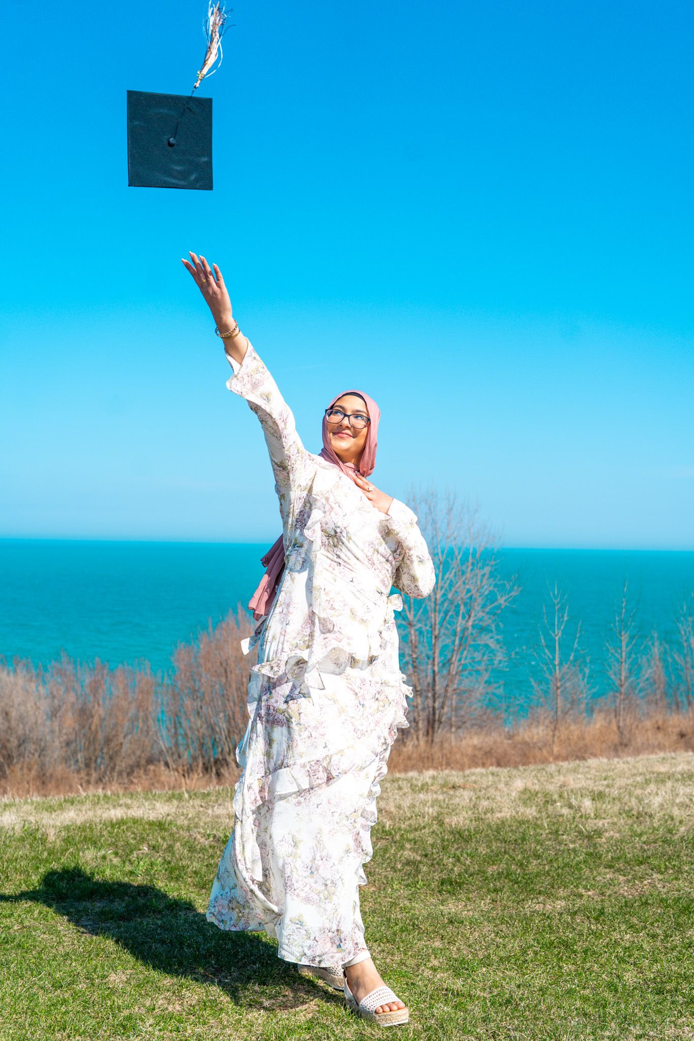 Graduate in white dress twirling joyfully on hilltop overlooking ocean view.