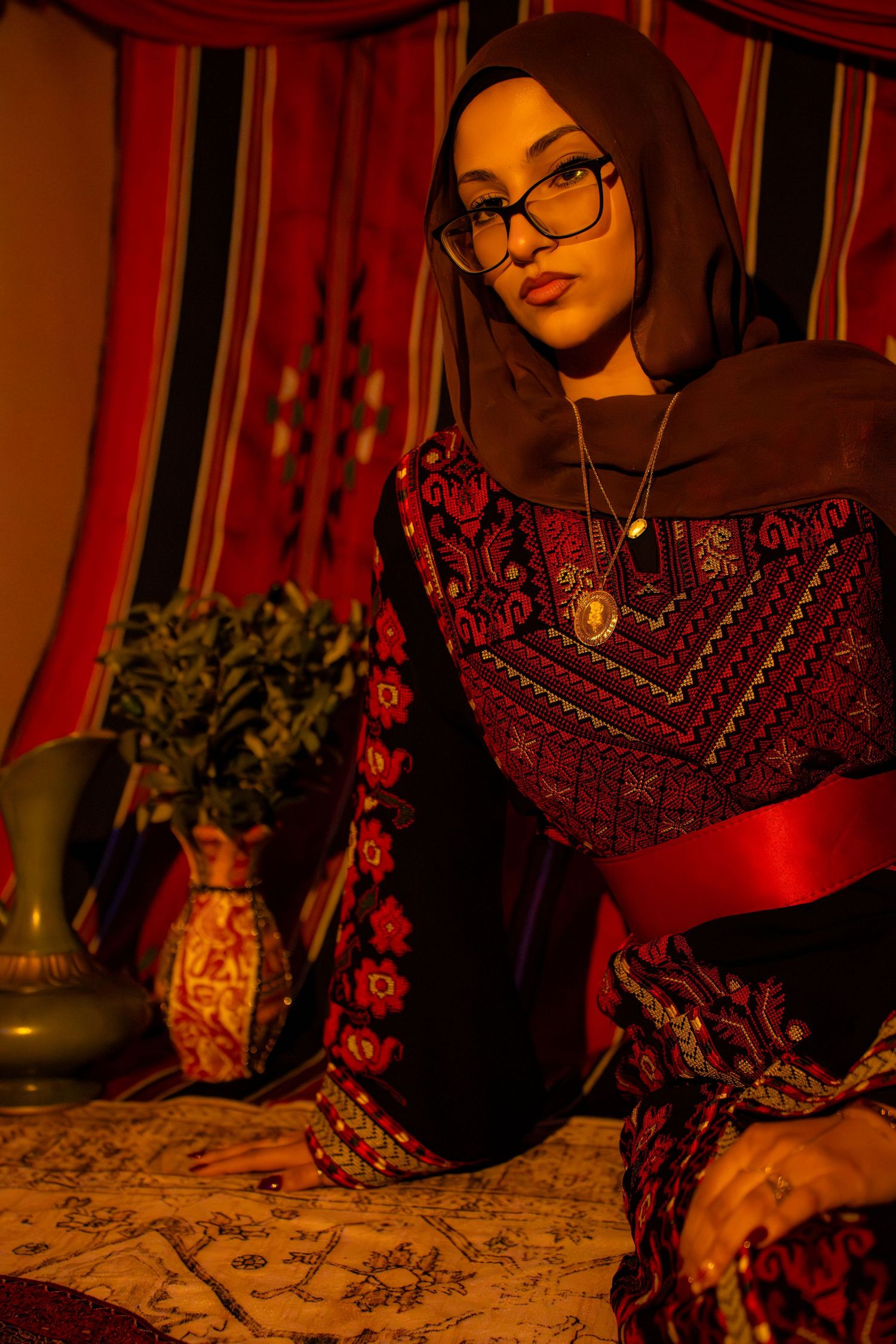 Moody portrait in warm lighting with red and black traditional dress, glasses, and decorative elements in background.