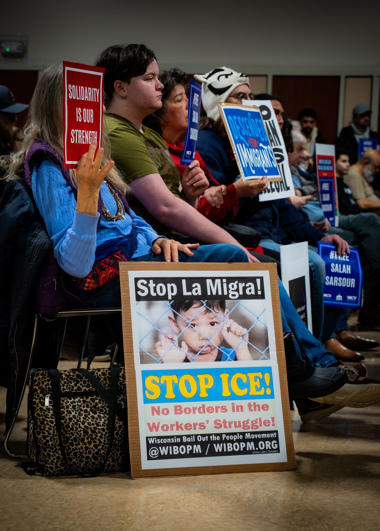 Protesters at rally holding signs reading Stop ICE, Stop La Migra, and Solidarity Is Our Strength at indoor demonstration.
