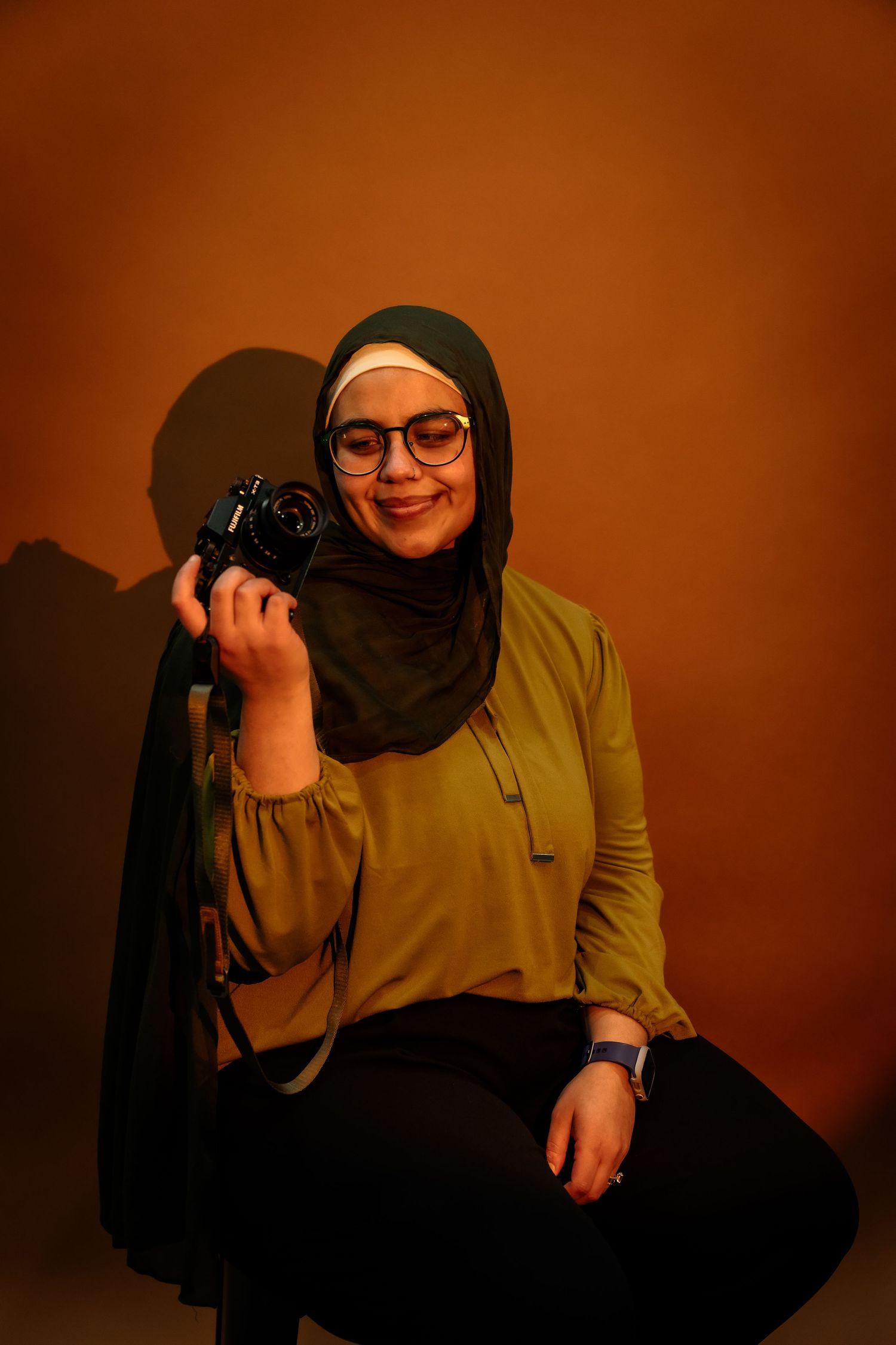 A photographer in a hijab and olive green top poses with a camera against a warm brown background in three shots.