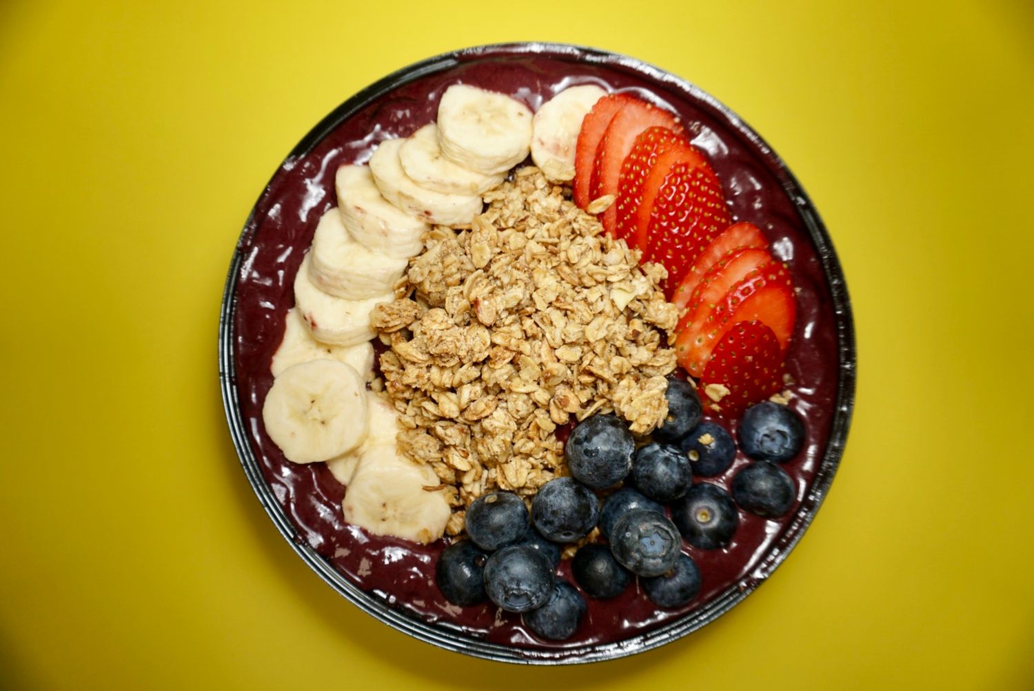 Acai bowl topped with fresh blueberries, bananas, granola and strawberries on yellow background.