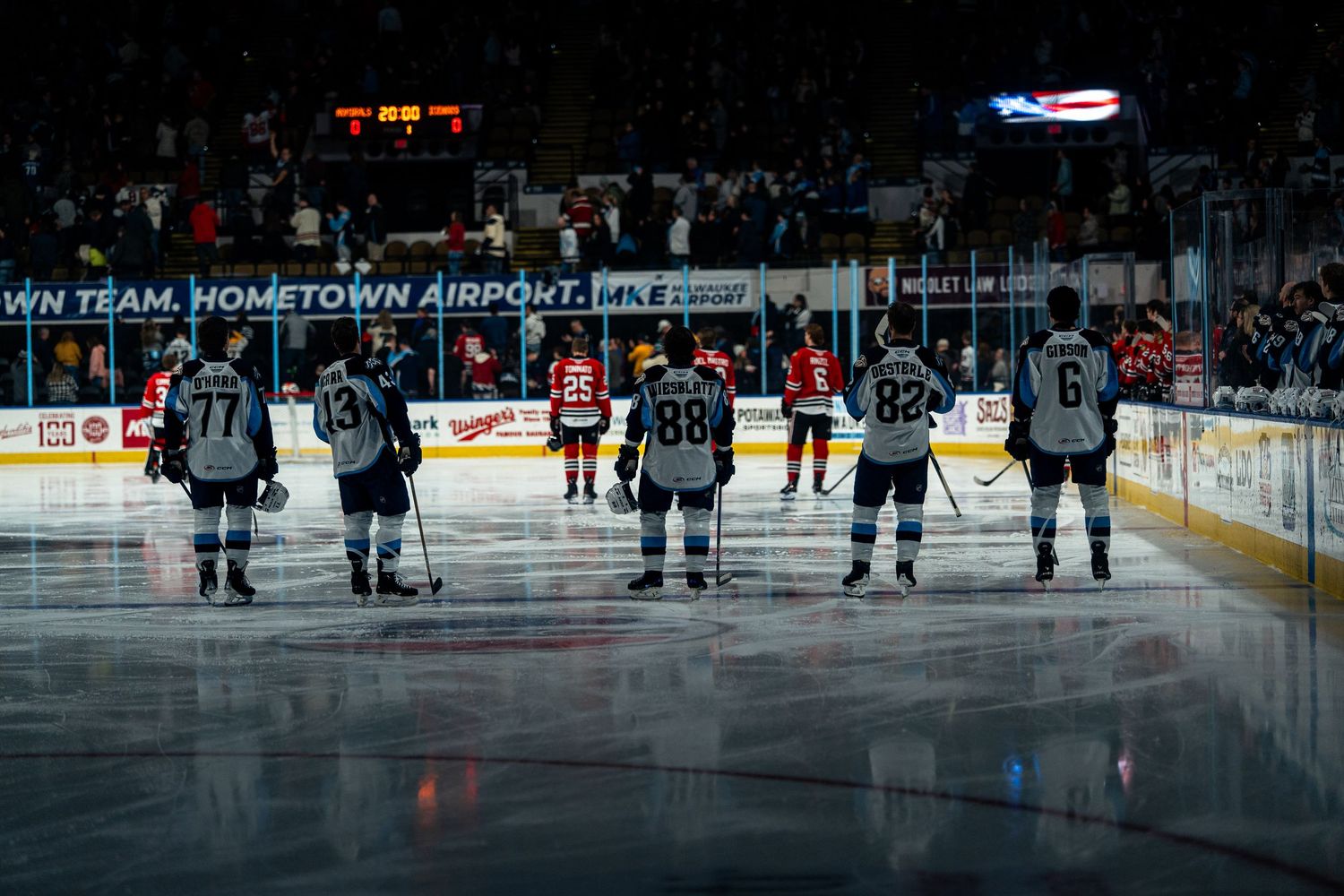 Hockey players line up on ice during pre-game ceremony with dark arena lighting.