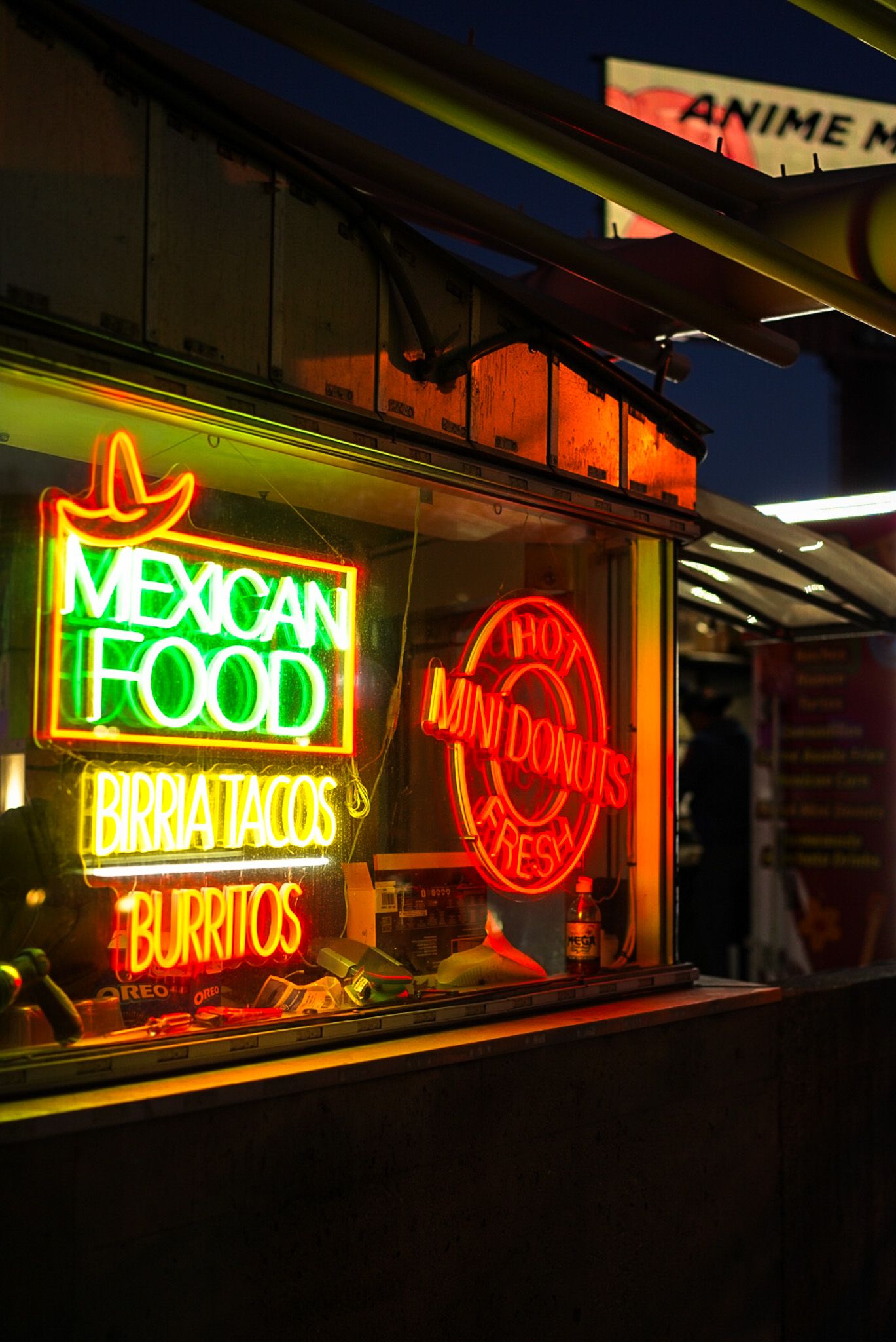 Colorful neon signs advertising Mexican food and burritos illuminate a dark storefront window.