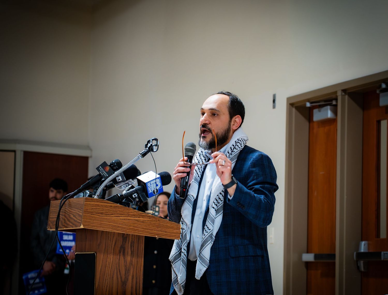 Man in suit with keffiyeh scarf speaks passionately at podium with microphones during public address event.