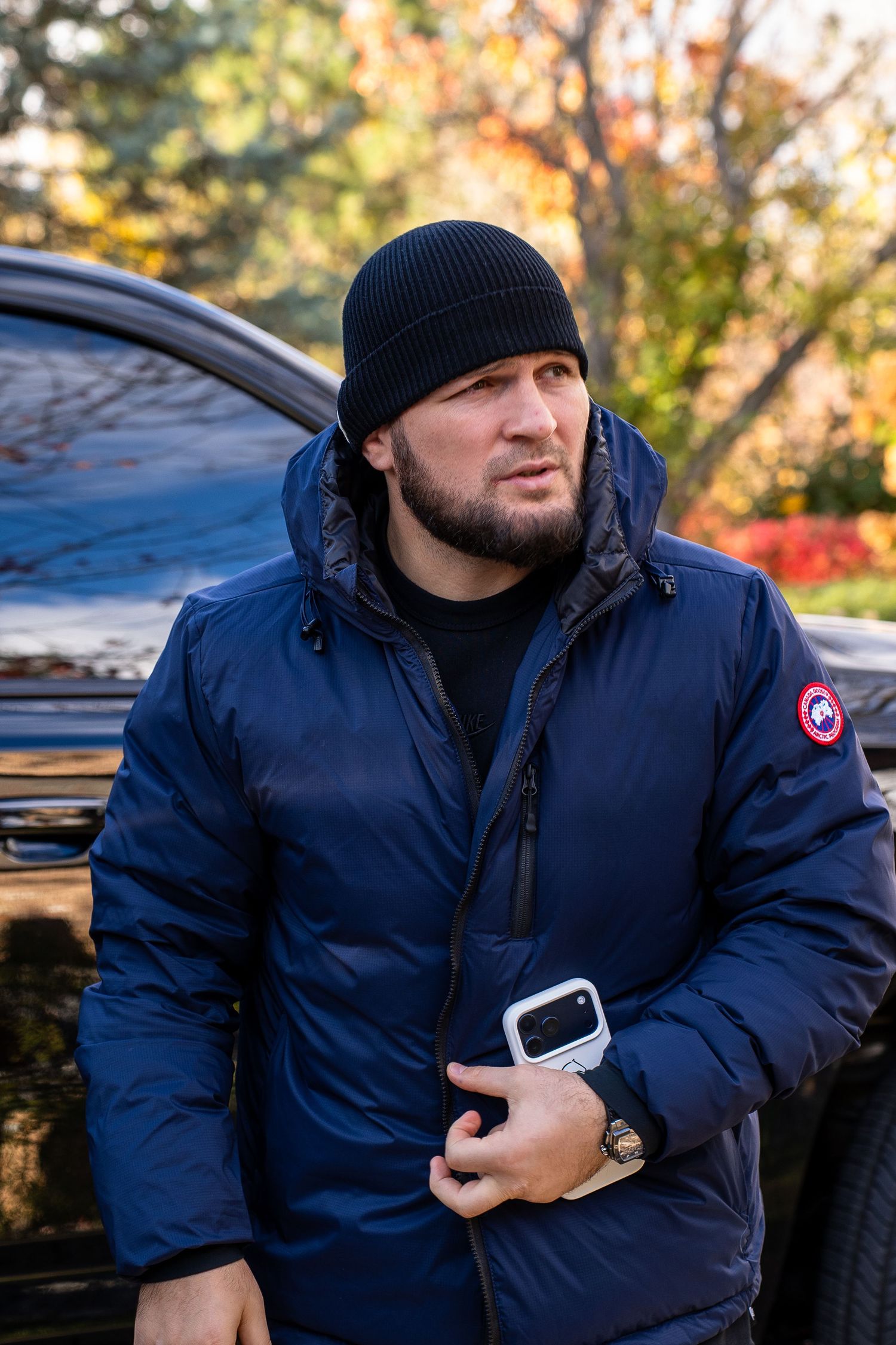 Someone wearing a navy Canada Goose winter jacket and black beanie stands outdoors on an autumn day.