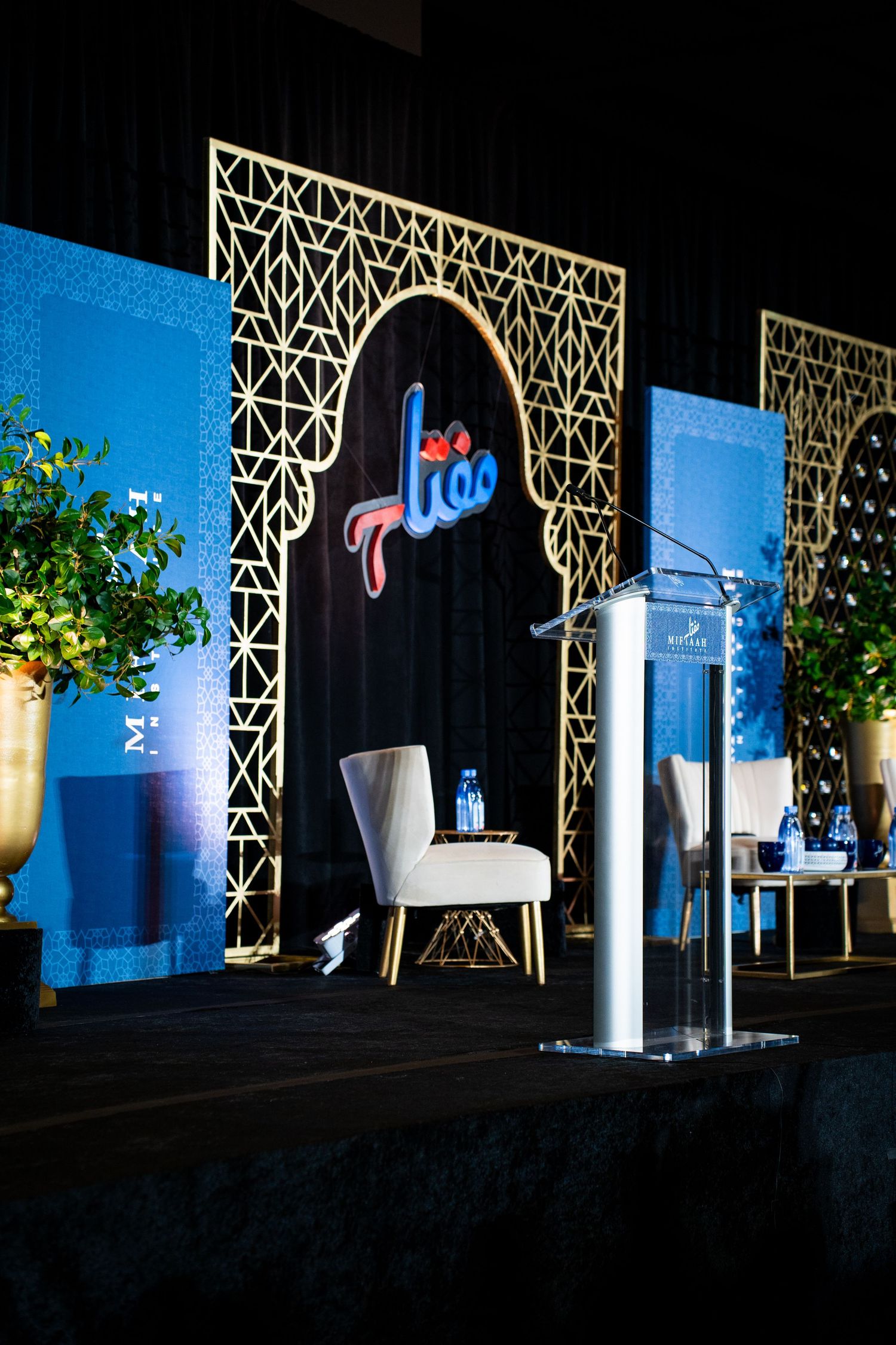 Stage setup with geometric lattice backdrop design illuminated in blue lighting with decorative plants.