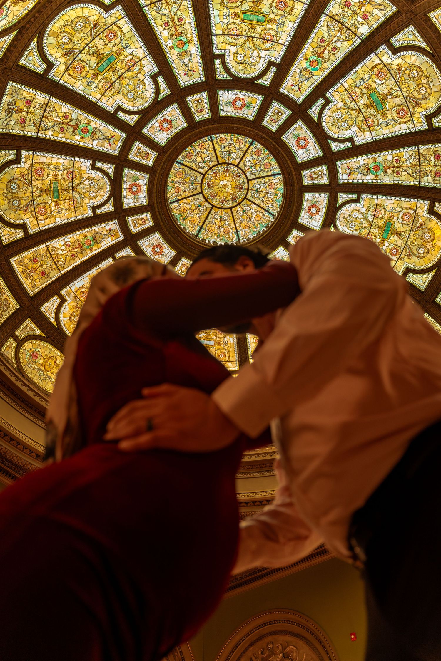 Two figures embrace beneath an ornate stained glass dome ceiling with golden floral patterns and geometric designs.