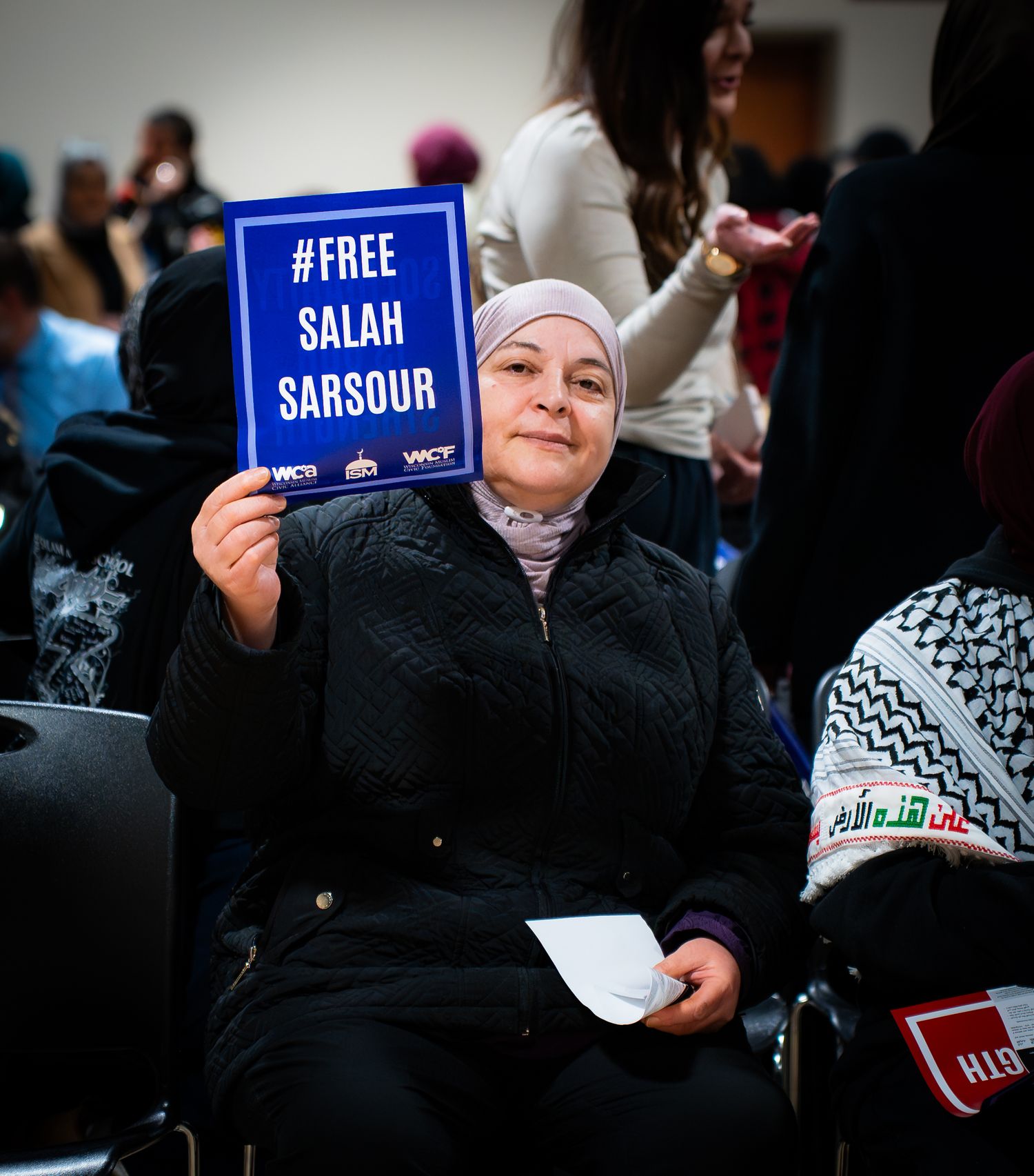 Woman in hijab holds a #Free Salah Sarsour sign at an indoor rally or meeting.