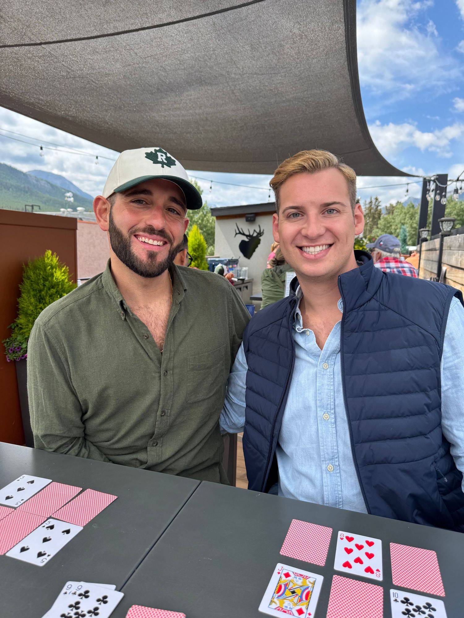 Two people sit at a table playing a card game outdoors on a sunny day with mountains visible in the background.