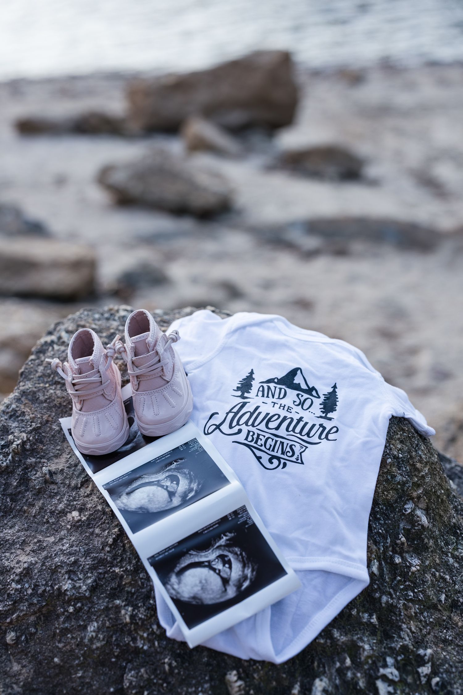 Baby announcement with ultrasound photos, pink shoes, and onesie reading And So The Adventure Begins on rocky beach.