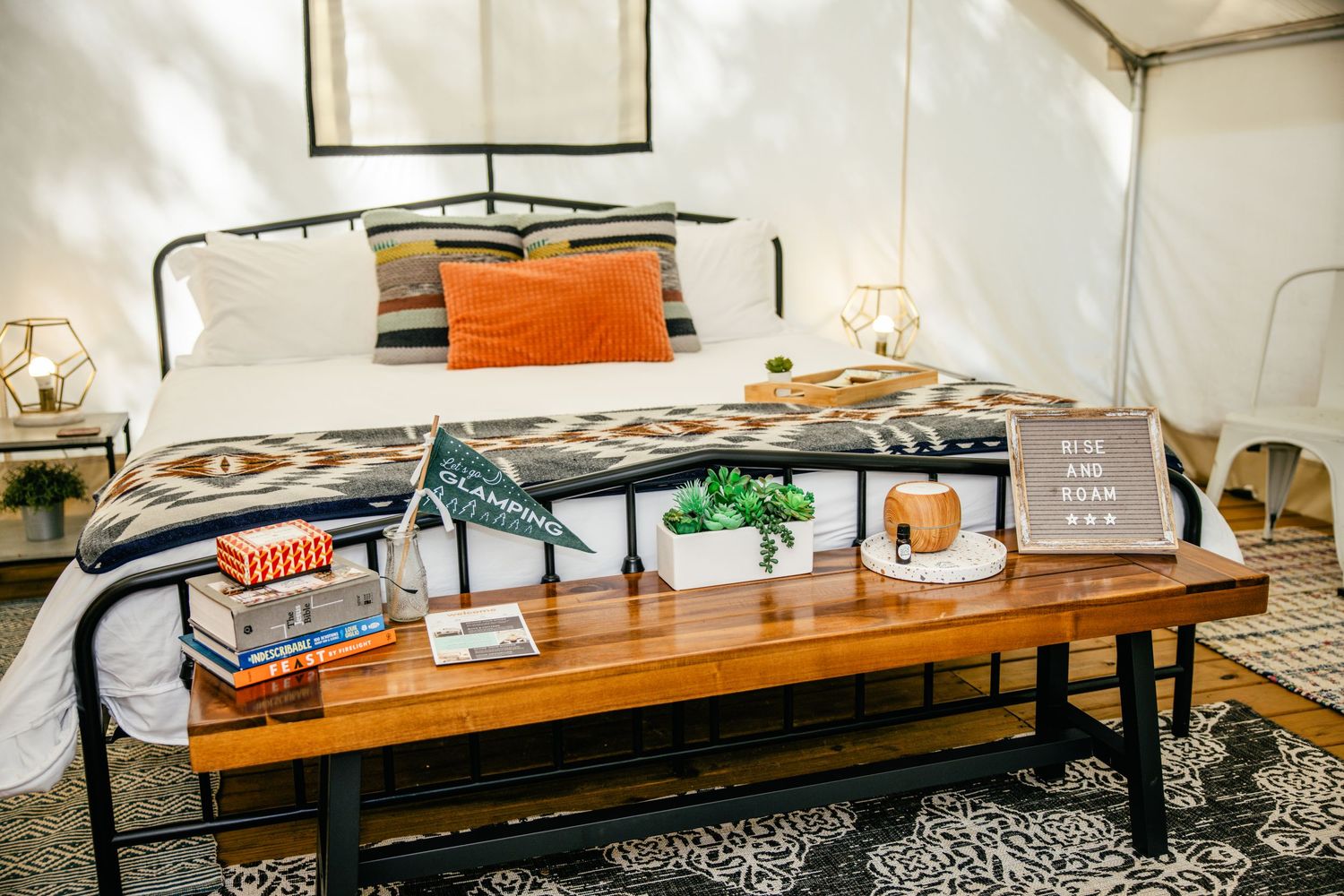 Cozy glamping tent bedroom with iron bed, orange pillows, Pendleton blanket, wooden bench, books, and succulents.