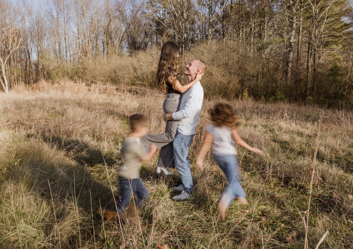 Father holds child while two kids play in a sunlit meadow surrounded by bare trees in autumn.