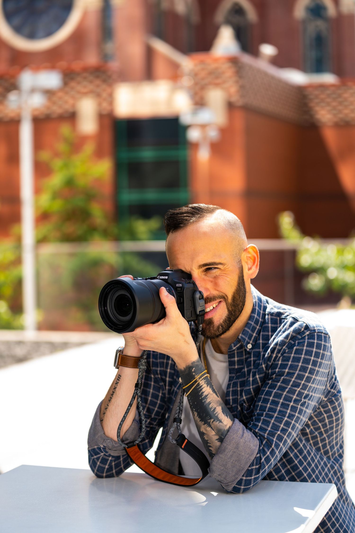 Professional photographer with tattoos and a plaid shirt smiling while holding a camera outdoors.