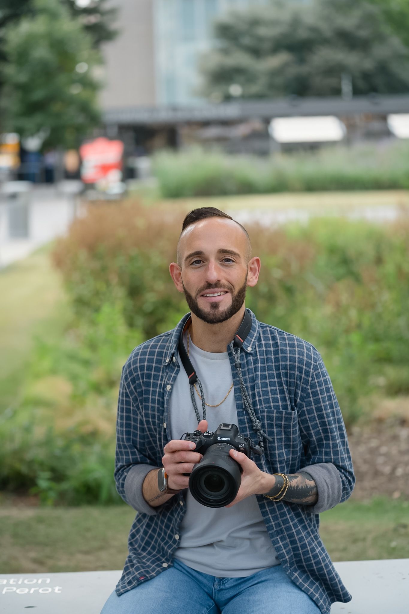 Smiling male photographer sitting outdoors holding a DSLR camera, wearing a plaid shirt and jeans.