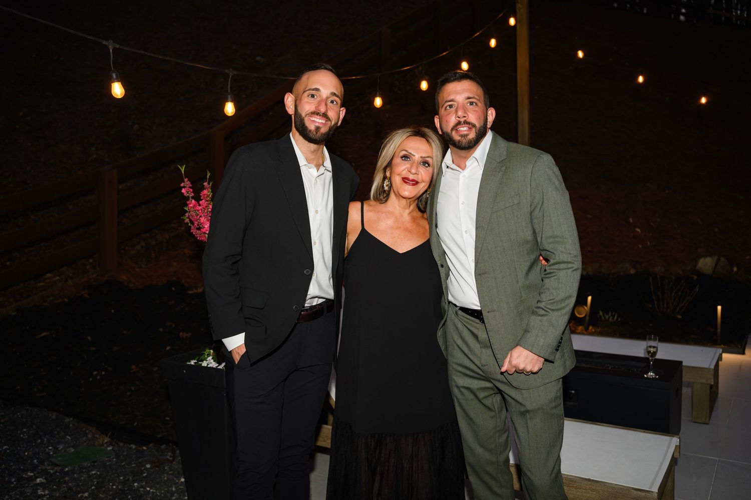 Three elegantly dressed people posing together at an evening event with string lights in the background.