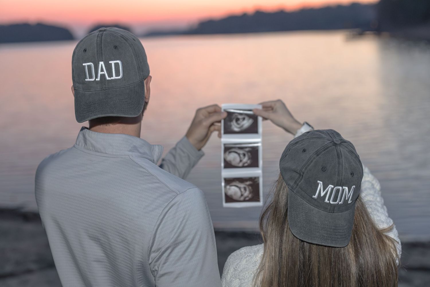 Couple wearing Dad and Mom hats viewing ultrasound photos by a lake at sunset, announcing pregnancy.