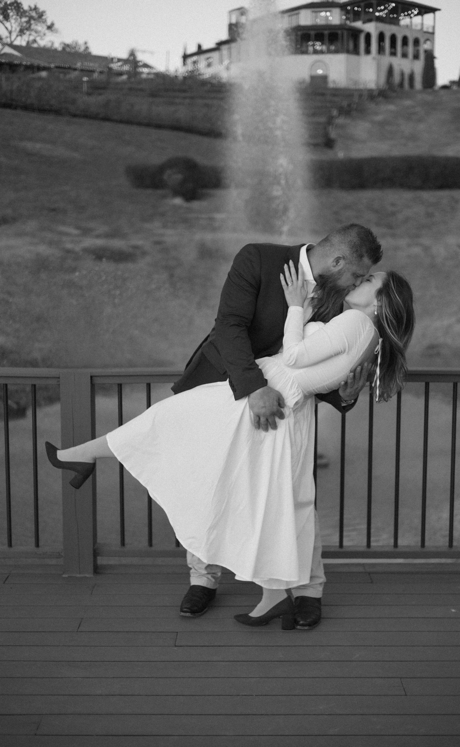 Black and white photo of a groom dipping and kissing his bride in a white dress on a deck with a fountain backdrop.