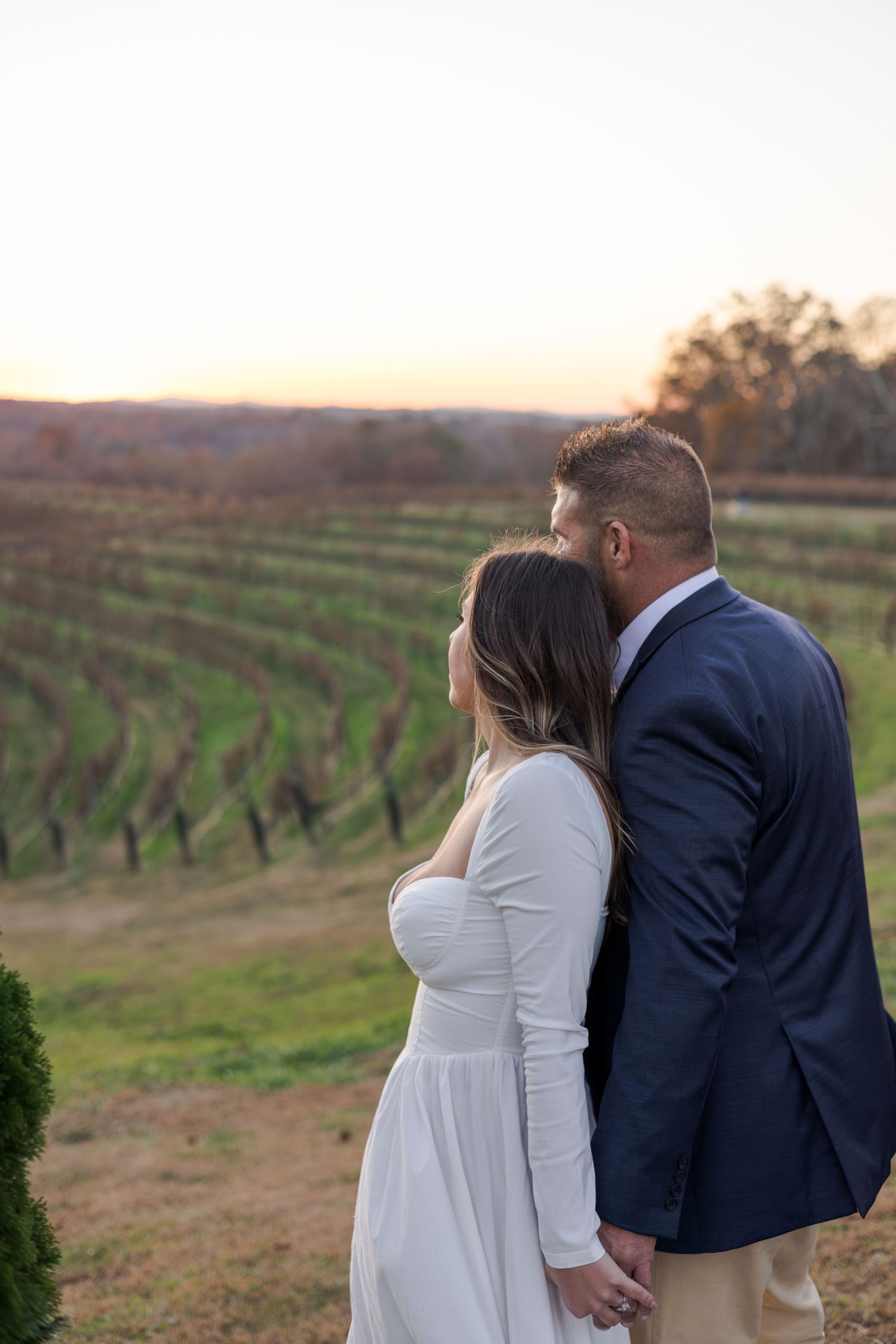 Couple holding hands at sunset overlooking a vineyard, man in navy blazer kissing woman in white dress on the cheek.