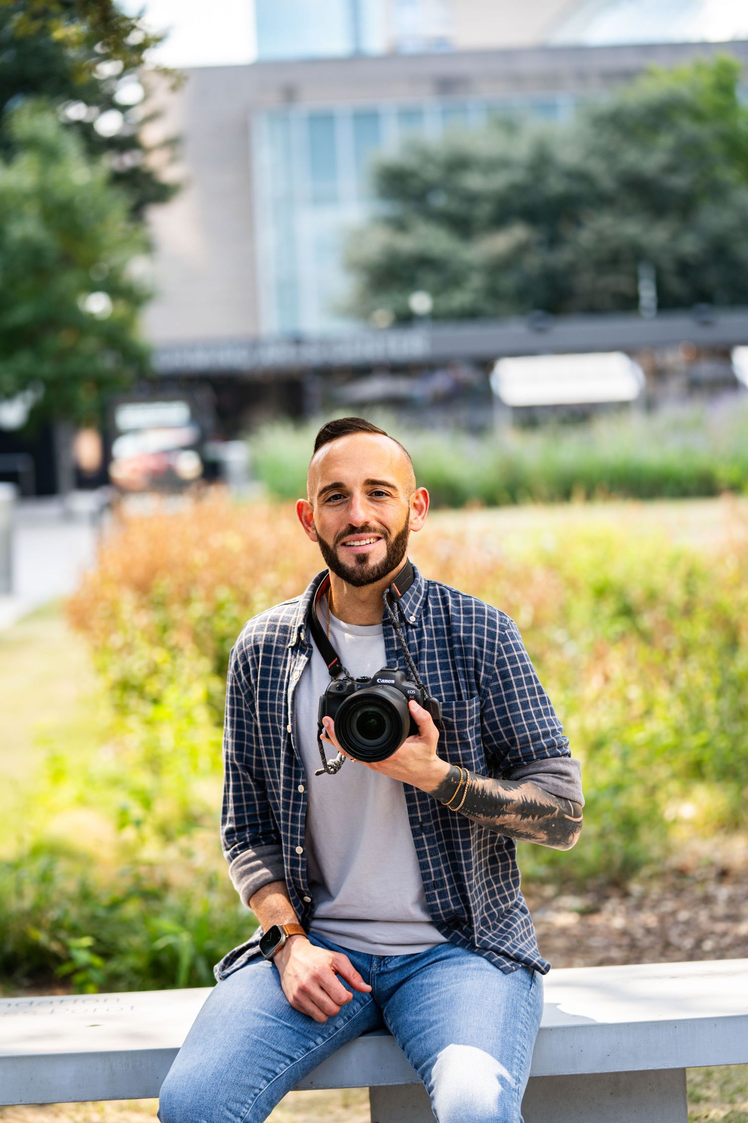 Professional photographer sitting on concrete ledge with camera in casual attire against blurred outdoor background.