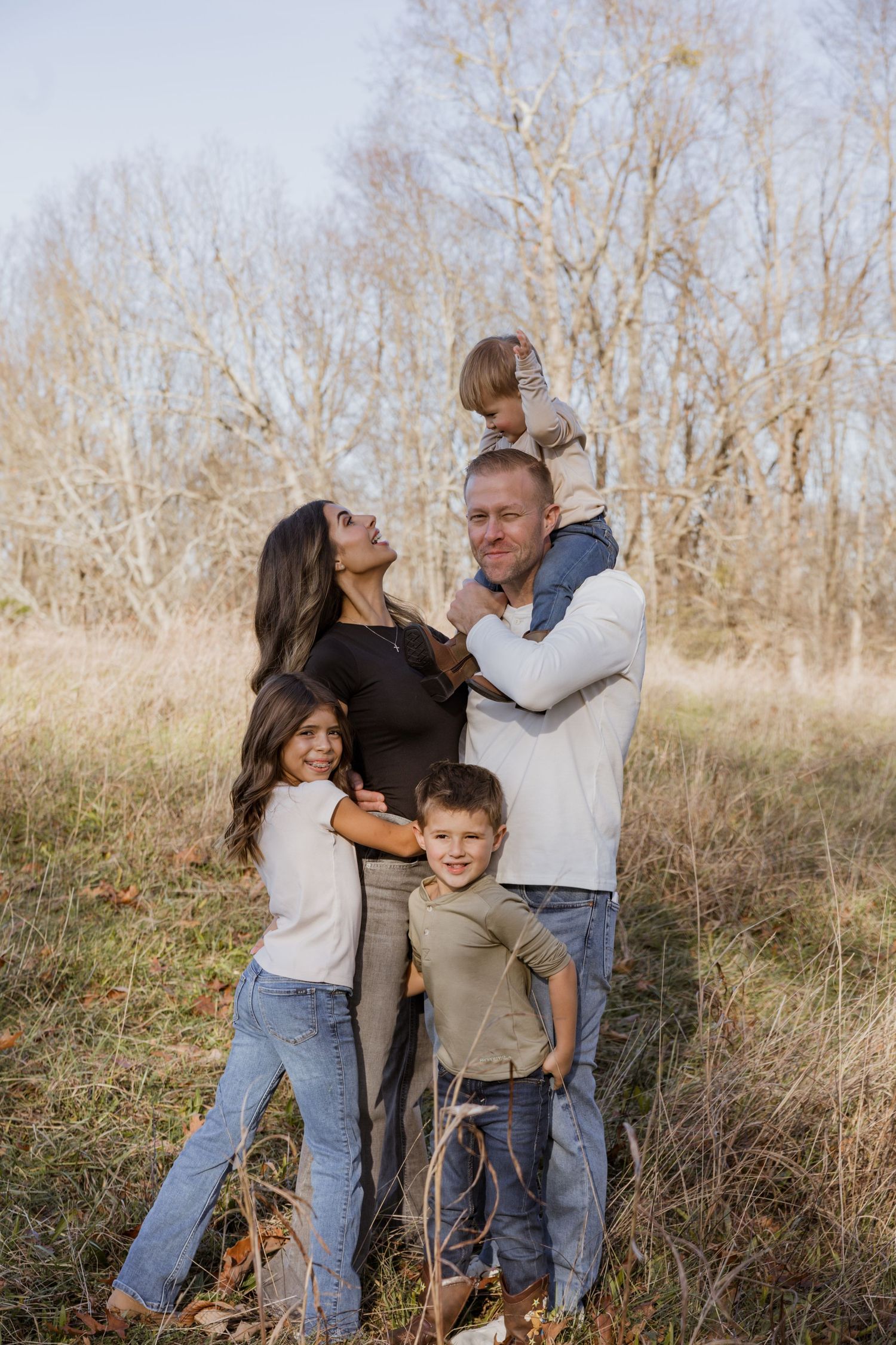 Family of five posing outdoors in a field with bare trees, parents and three children smiling in casual fall attire.