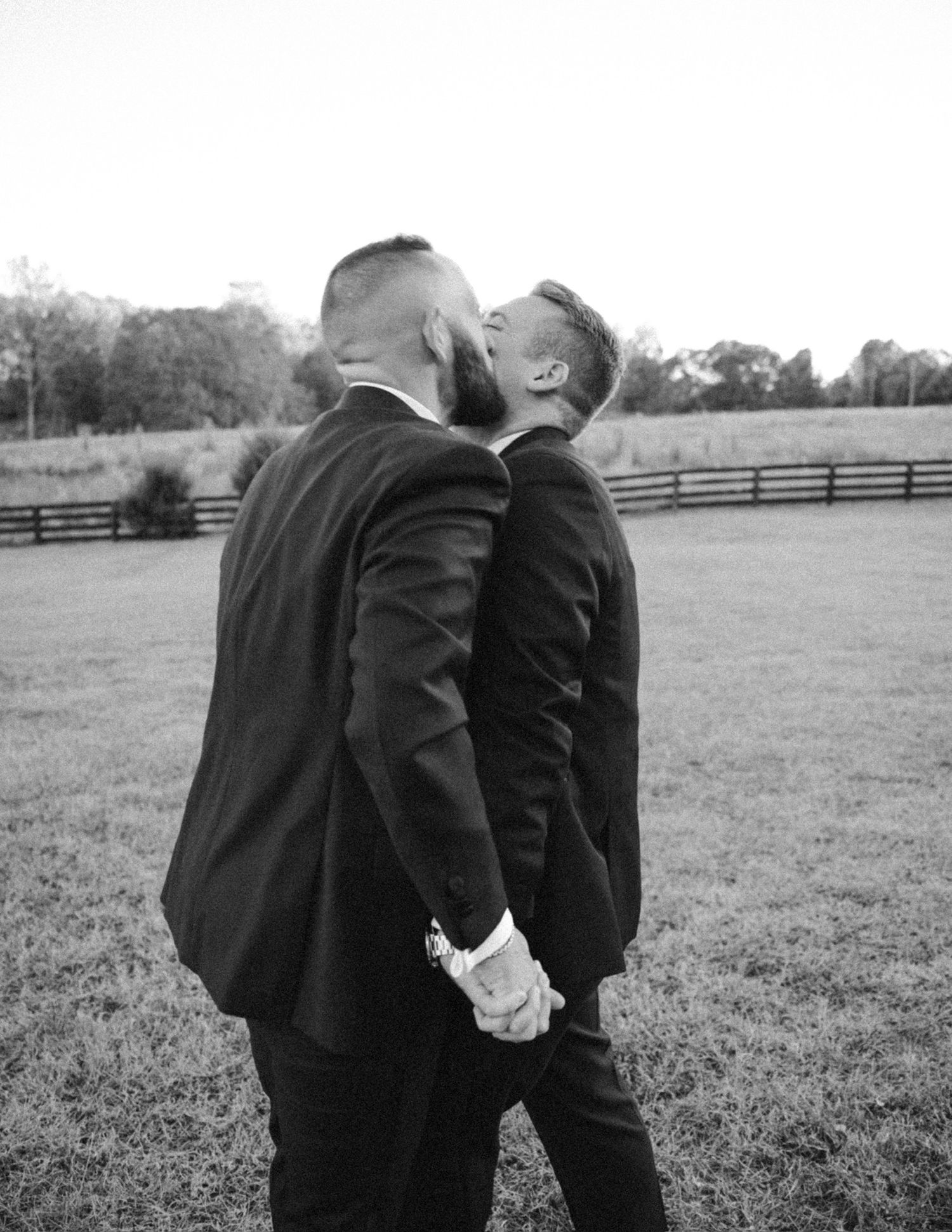 Two people in dark suits sharing an intimate moment in a field with wooden fences in the background.