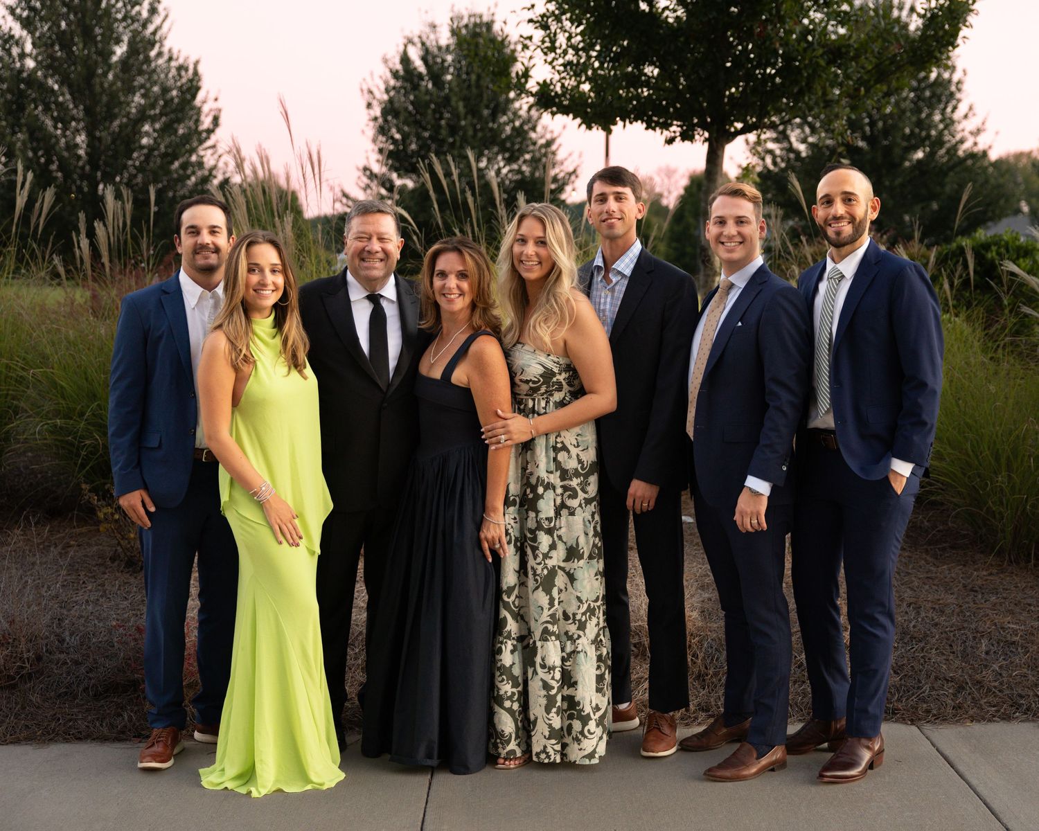 Group photo of wedding party members in formal attire outdoors at sunset.