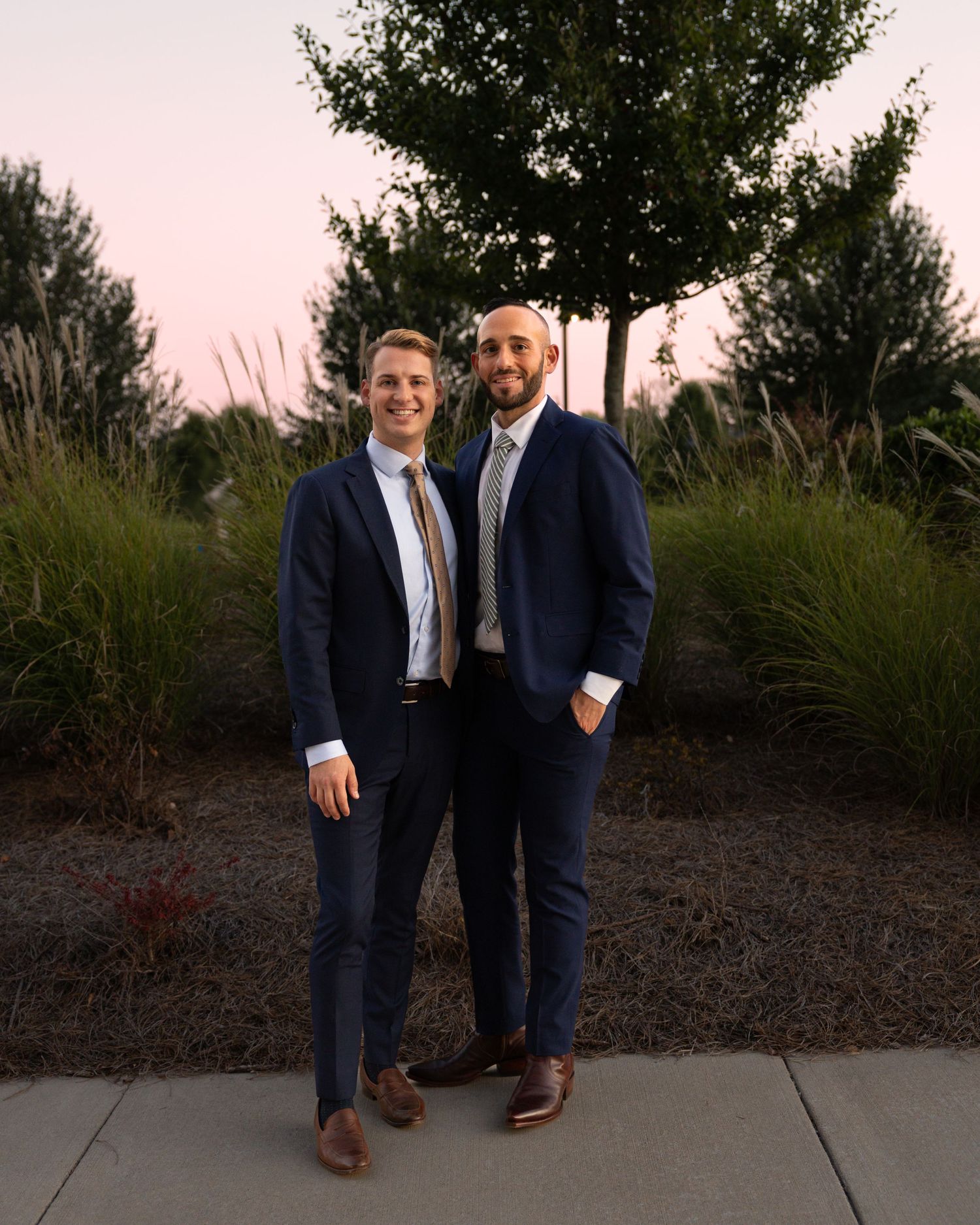 Two people in formal suits standing together outdoors at sunset.