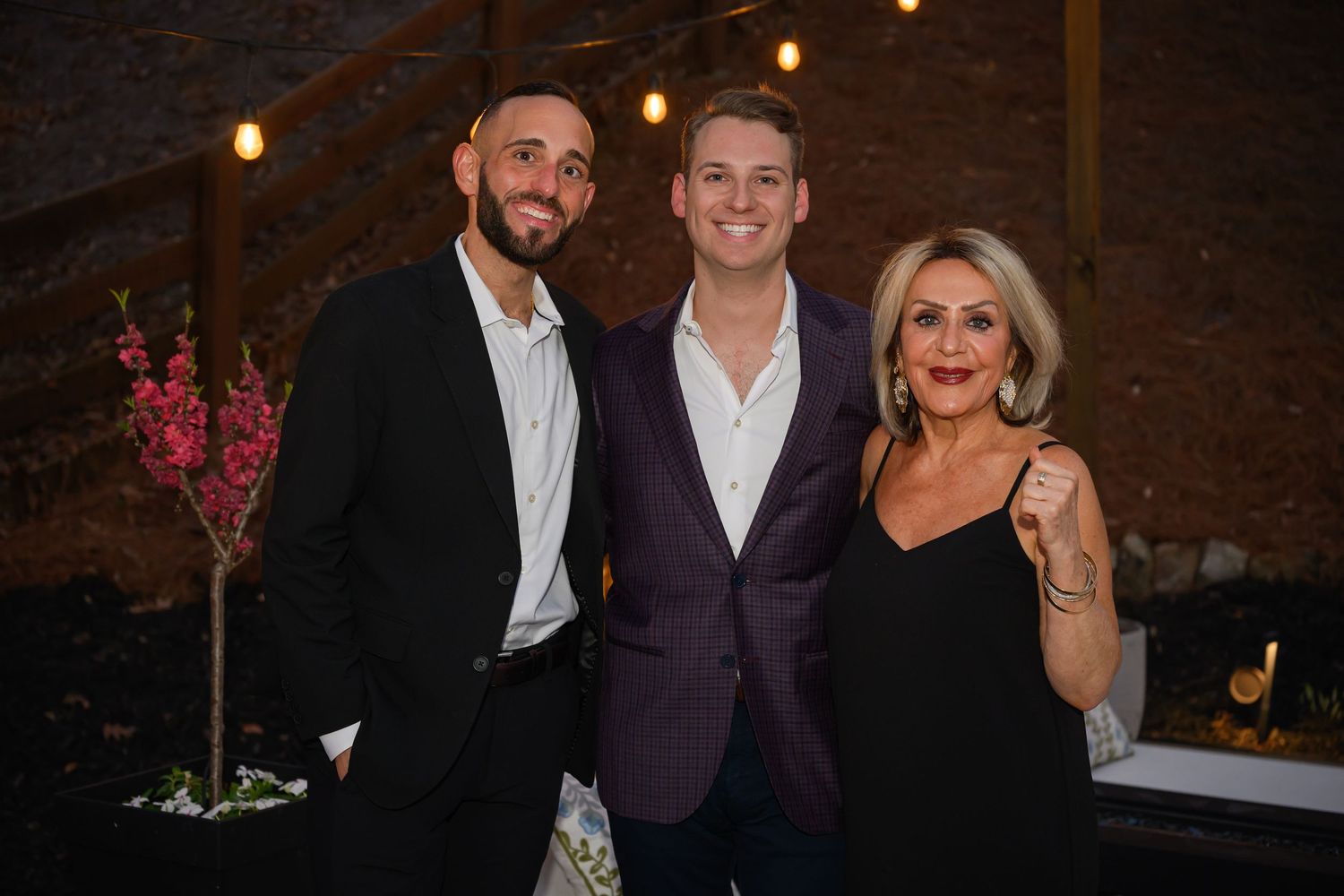 Three elegantly dressed people smiling together at an indoor evening event with warm string lights in the background.