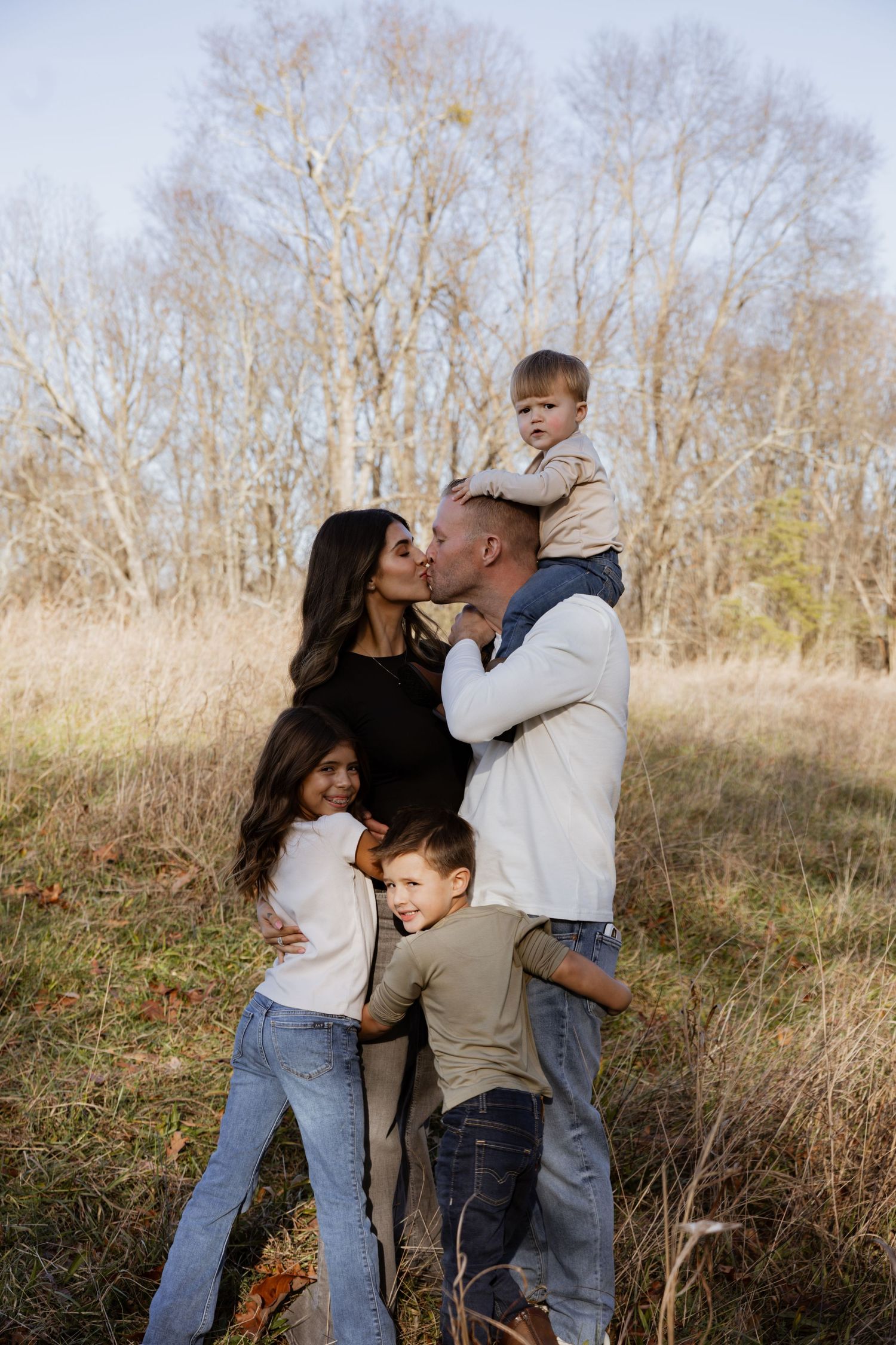 Family of five posing outdoors in autumn field, parents kissing while children hug them, bare trees in background.