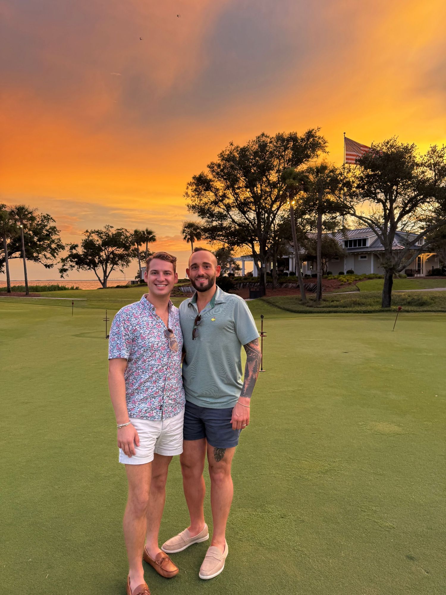 Scenic golf course at sunset with silhouetted trees and vibrant orange sky casting warm light across the green fairway.