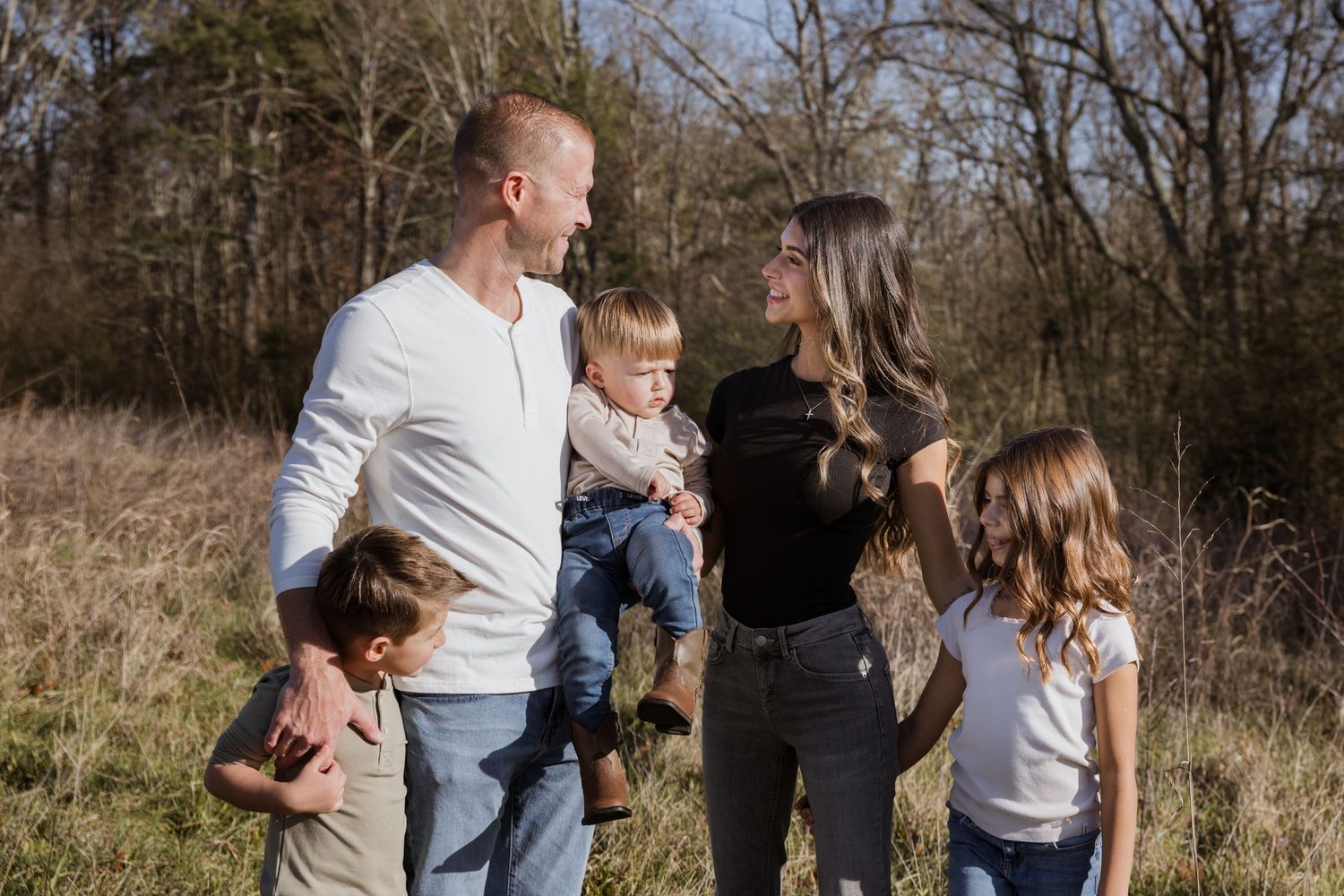 Family of five posing outdoors in a sunny field with trees, parents holding young children in a candid portrait.