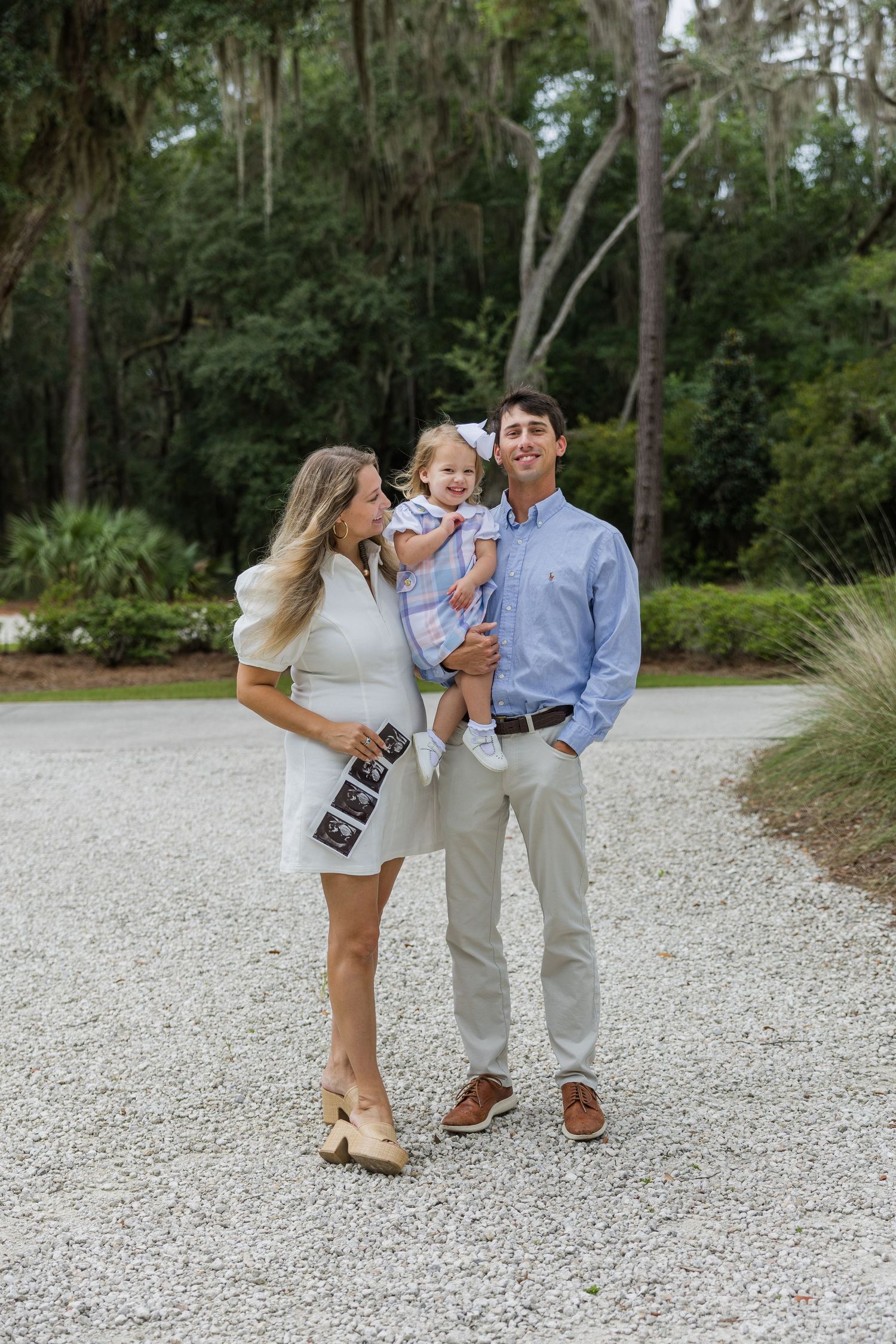 Happy family pregnancy announcement on gravel path, woman in white dress holding ultrasound photos, man carrying toddler girl.