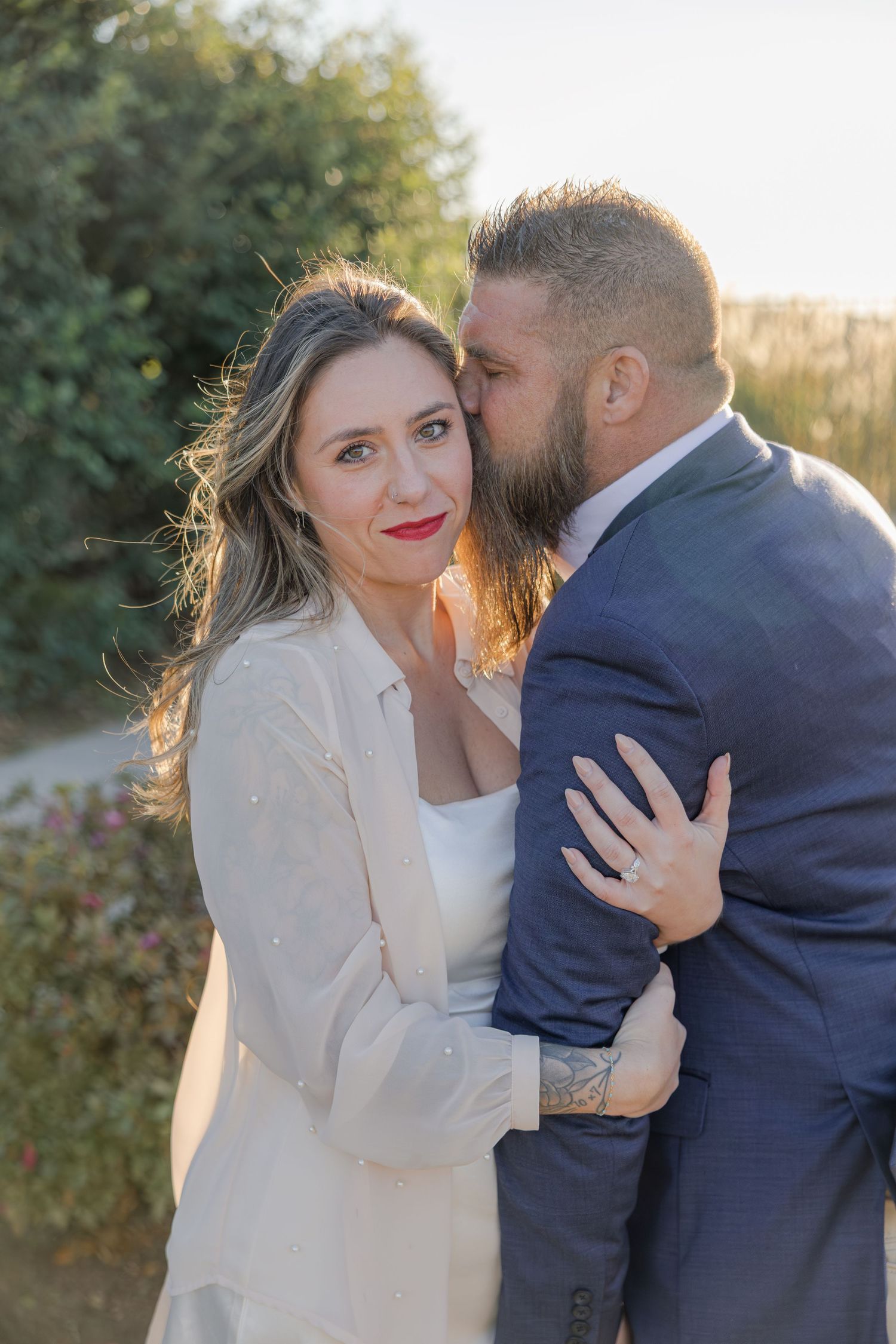 Couple embracing at golden hour, bride in white coat with red lips, groom in navy suit kissing her cheek outdoors.