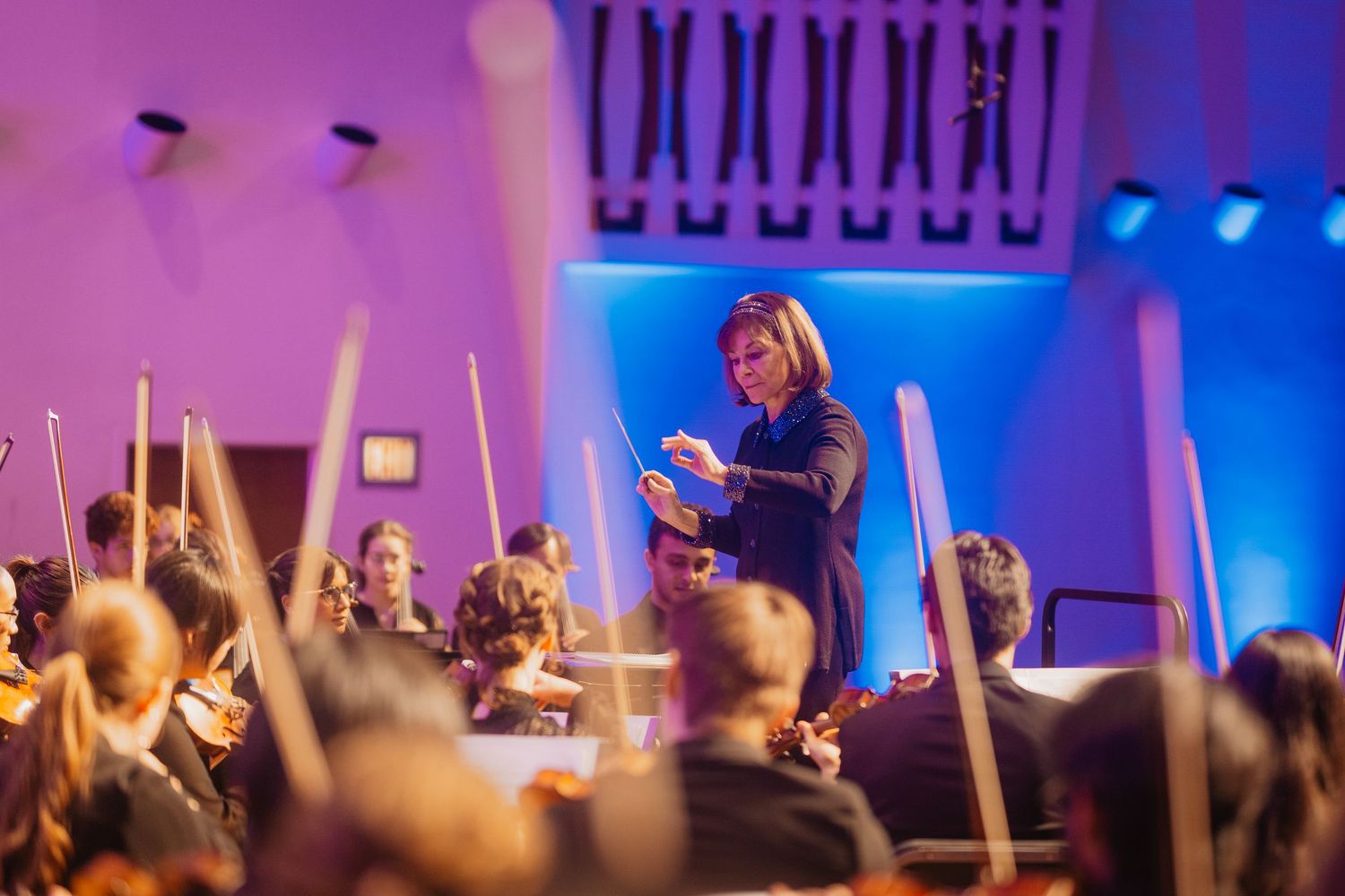 A female conductor leads an orchestra with passion under colorful blue and purple stage lighting.