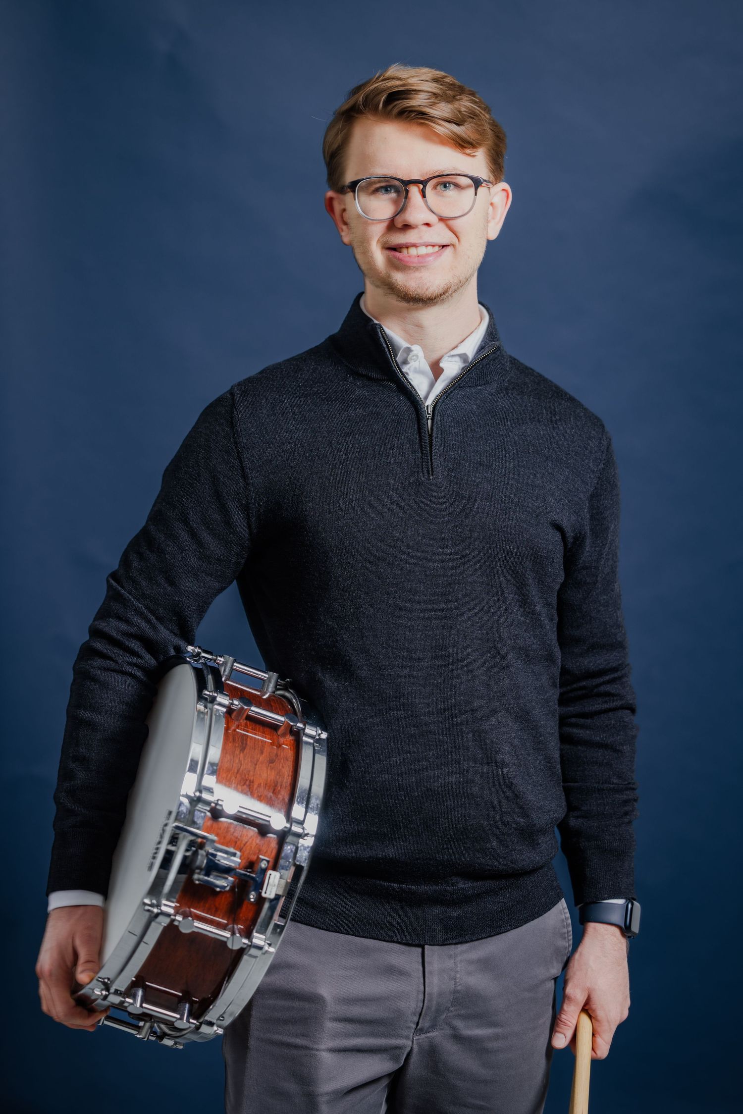 Smiling male percussionist holding a snare drum and drumstick, wearing a gray sweater on a blue background.