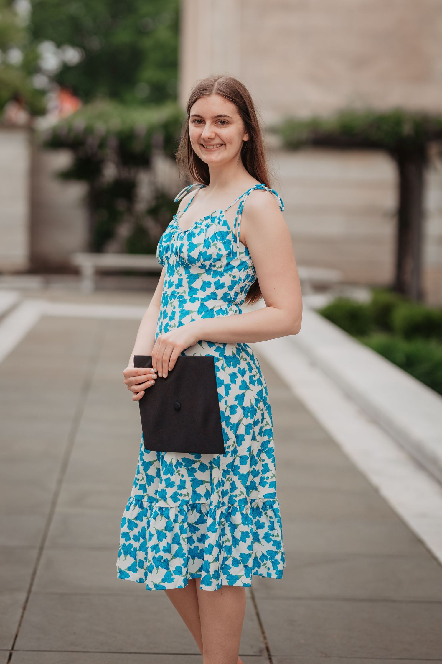 Smiling young woman in blue floral dress holding black graduation cap outdoors.