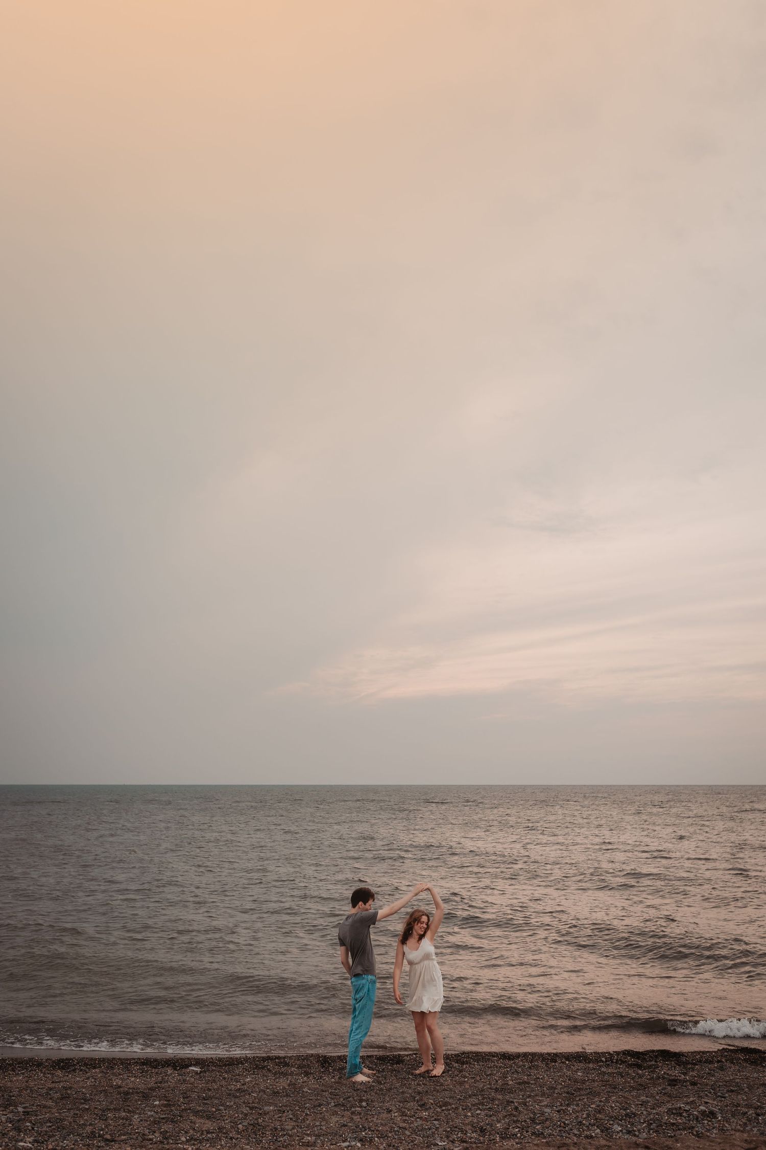 Romantic couple dancing on rocky beach shore at sunset with calm ocean horizon behind them.