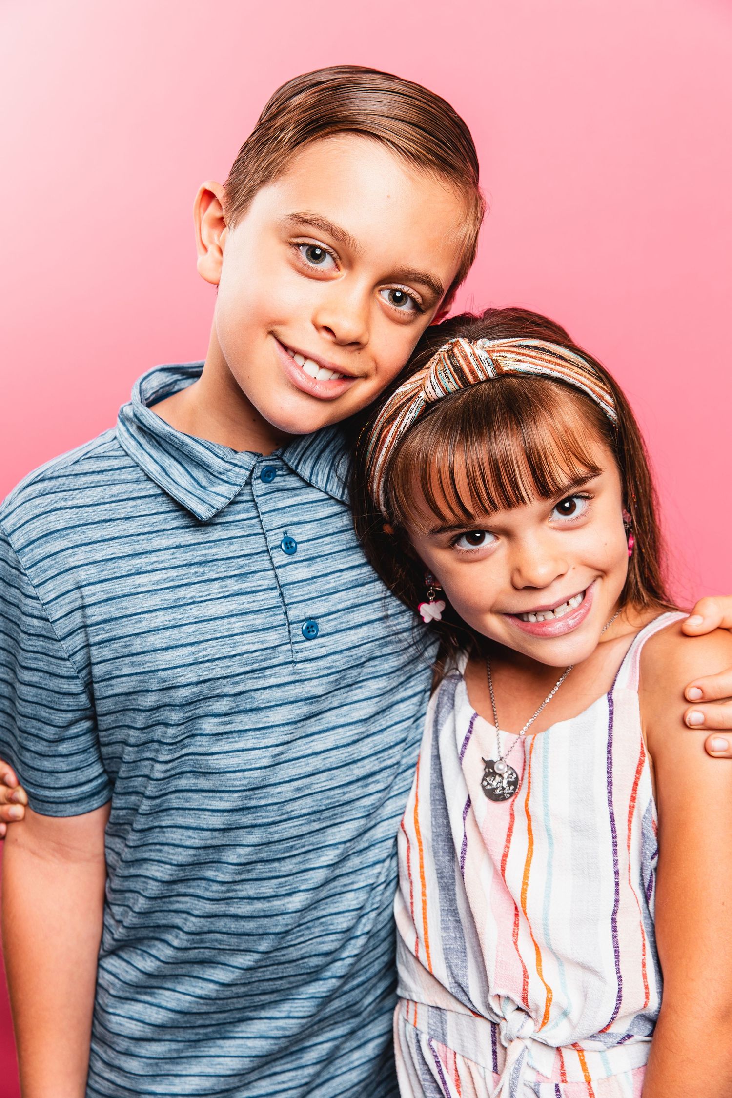 A smiling boy and girl pose together against a bright pink studio background.