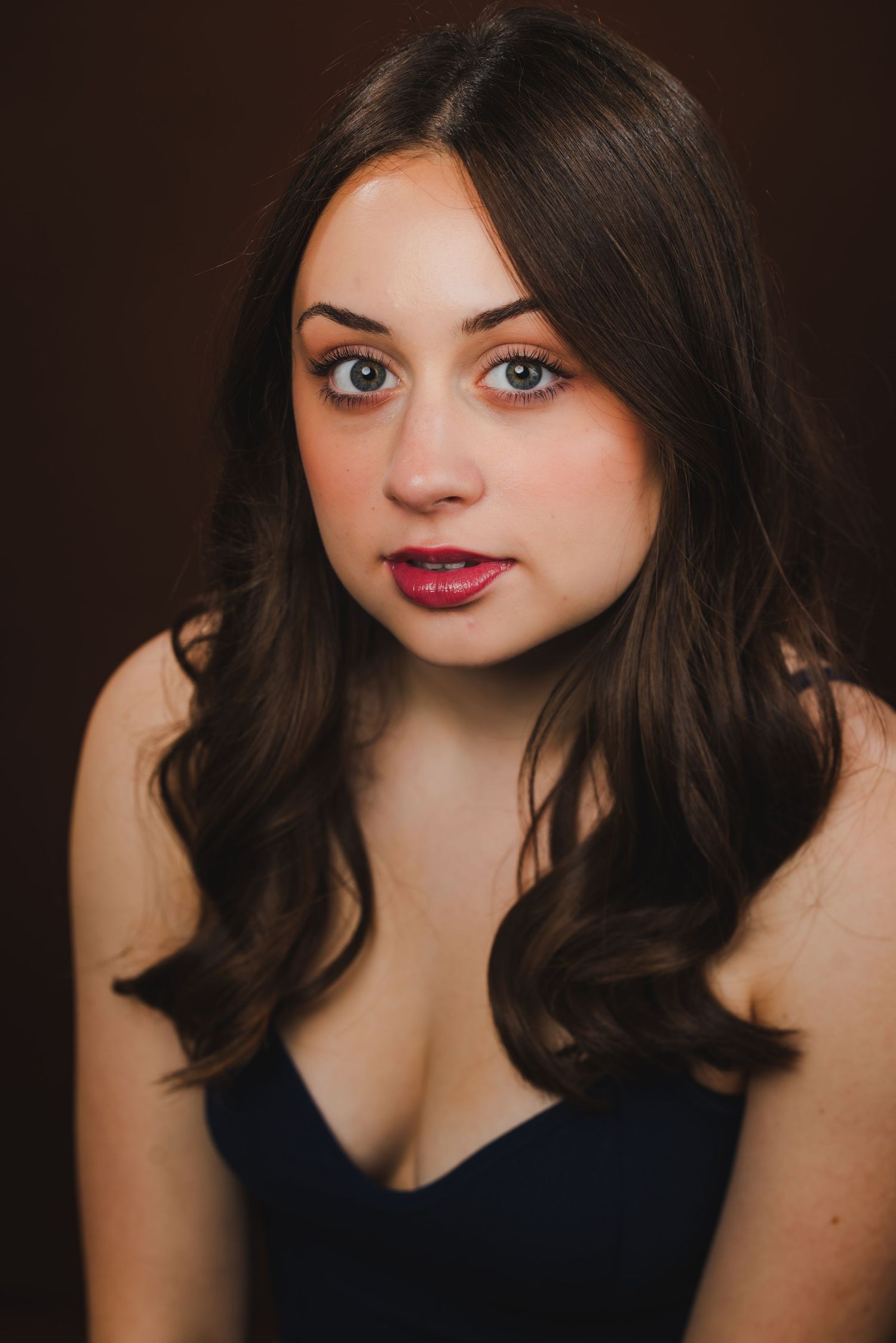 A young woman with long dark hair wearing a navy top with red lipstick poses against a brown background.