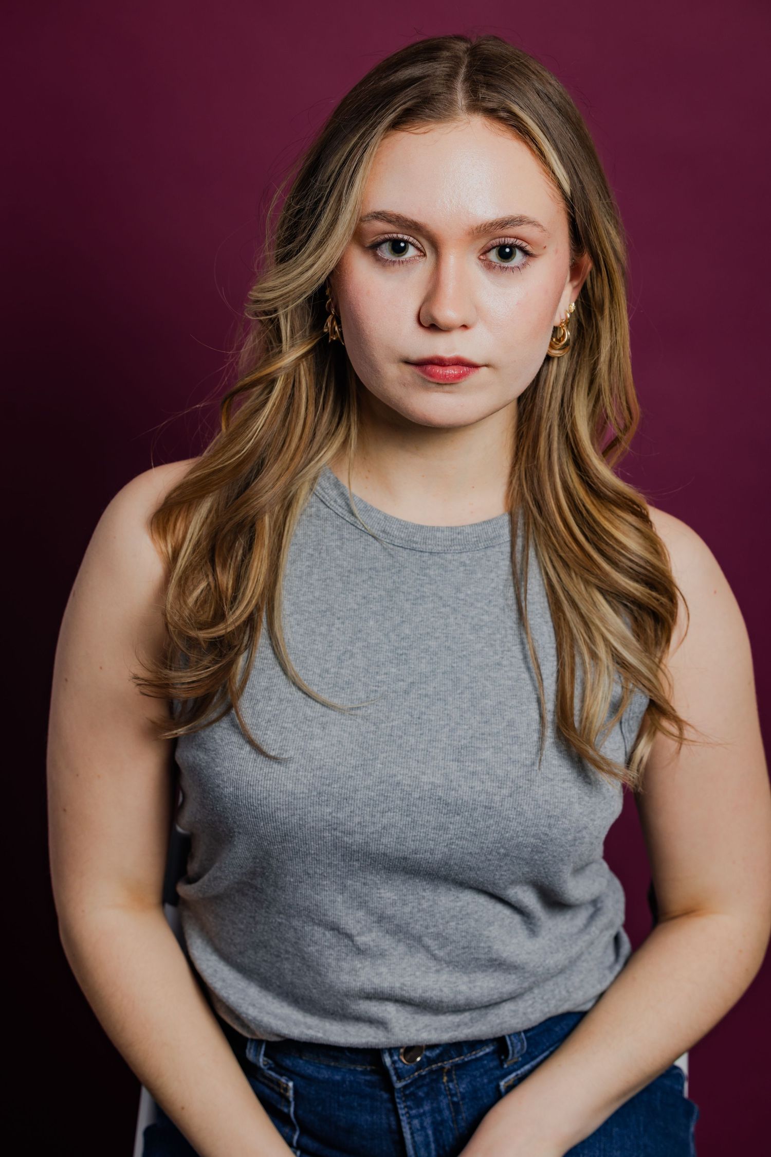 Young blonde woman in a gray tank top and jeans poses against a dark burgundy background.
