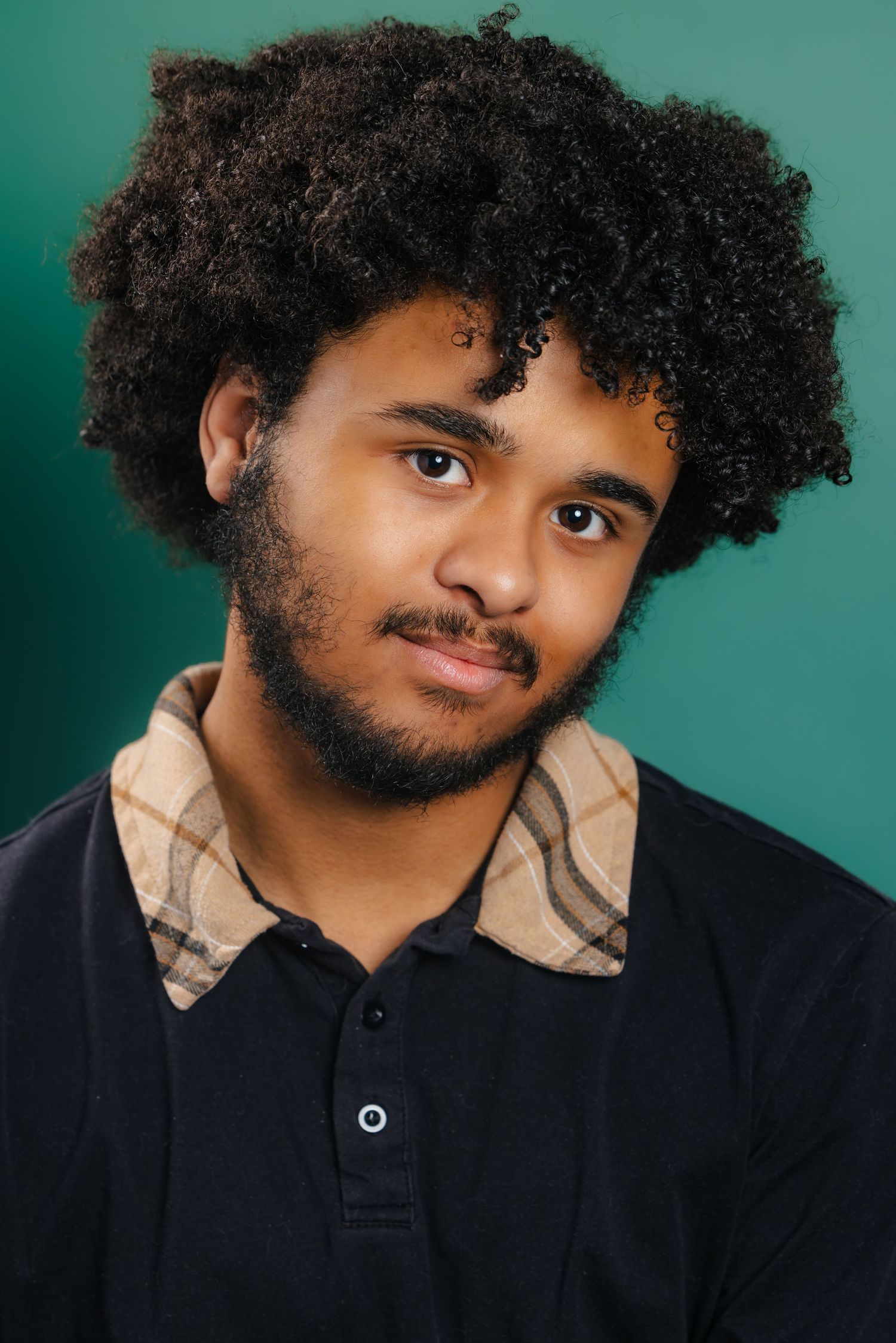 Young man with curly afro hair and beard wearing black polo shirt, teal background portrait.