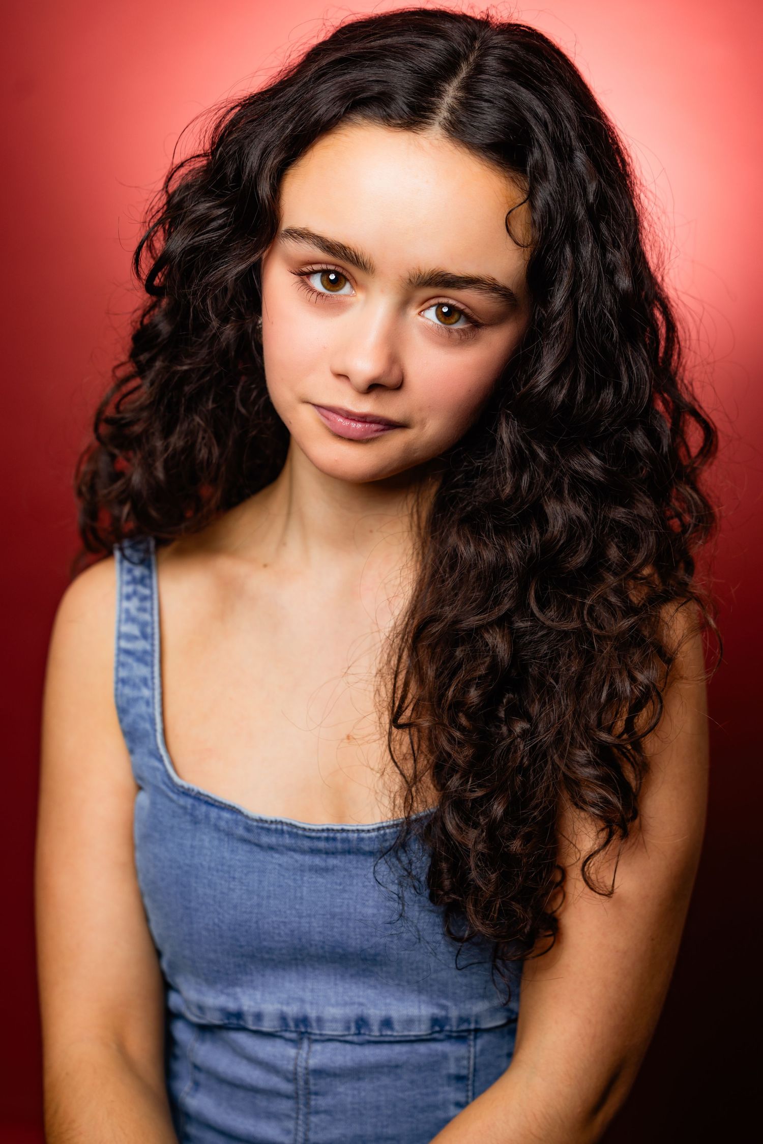 Young woman with long dark curly hair wearing a denim top poses for a professional headshot on red background.