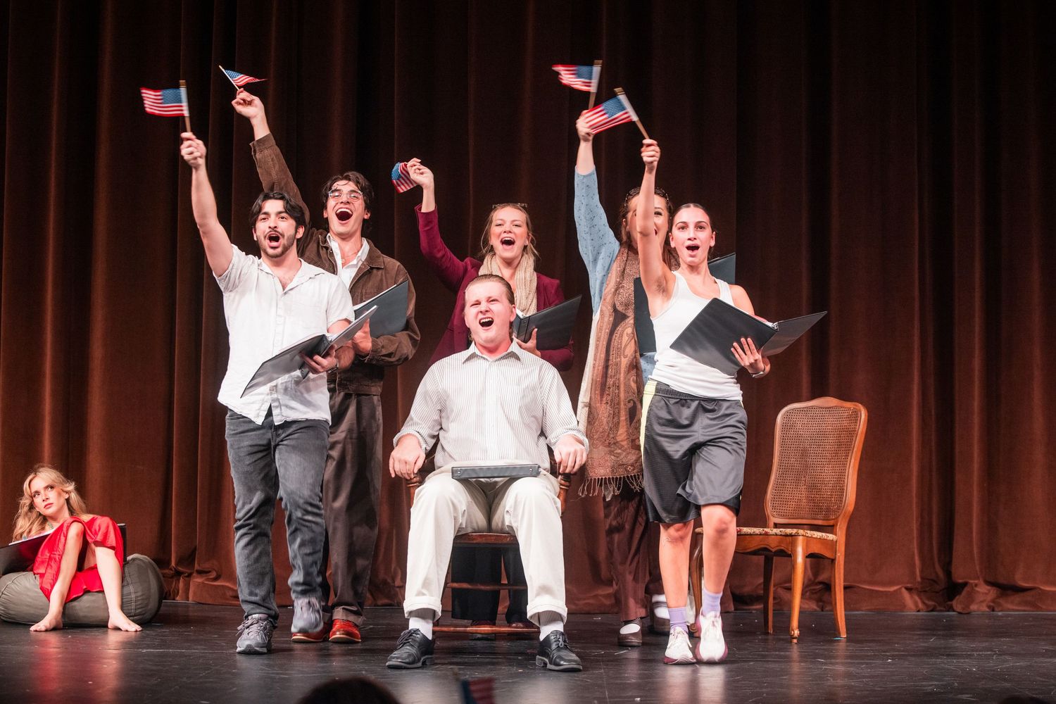 Energetic theater cast celebrating on stage waving small flags during a musical performance.