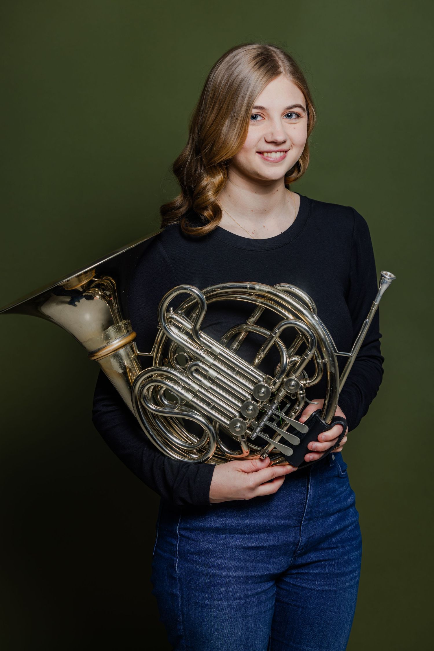 Young woman smiling and holding a French horn against a dark green background.