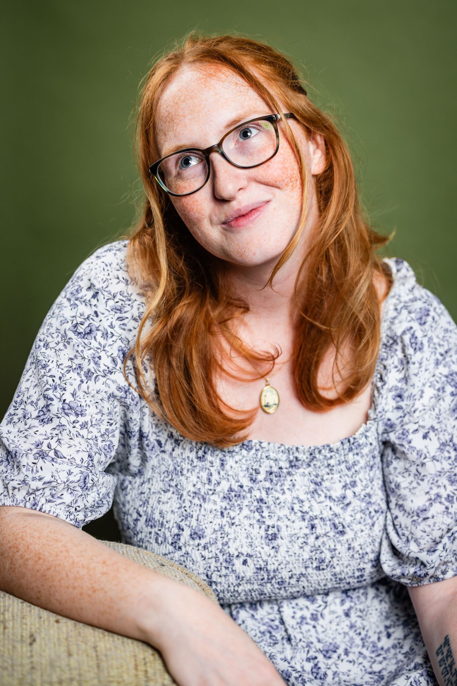 Young woman with red hair, freckles, and glasses wearing floral dress with cameo necklace on green background.