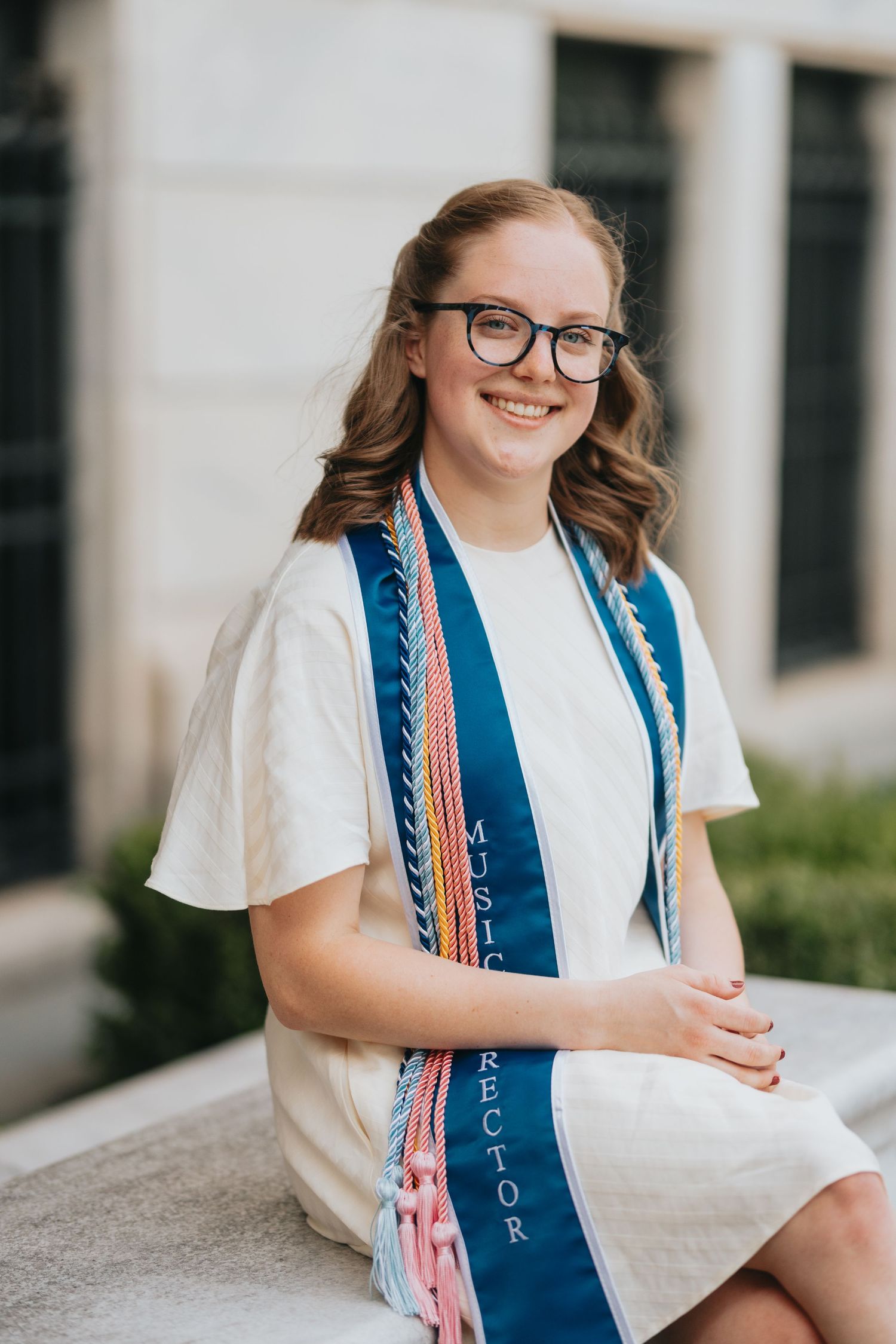 Smiling young woman with glasses wearing Music Director sash seated on stone ledge.