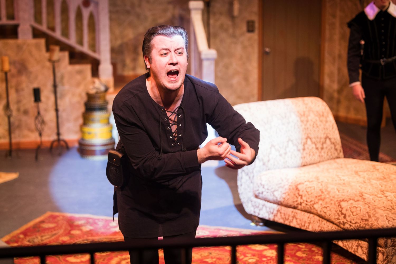 A dramatic actor in black clothing performs an intense scene leaning over a couch on stage.